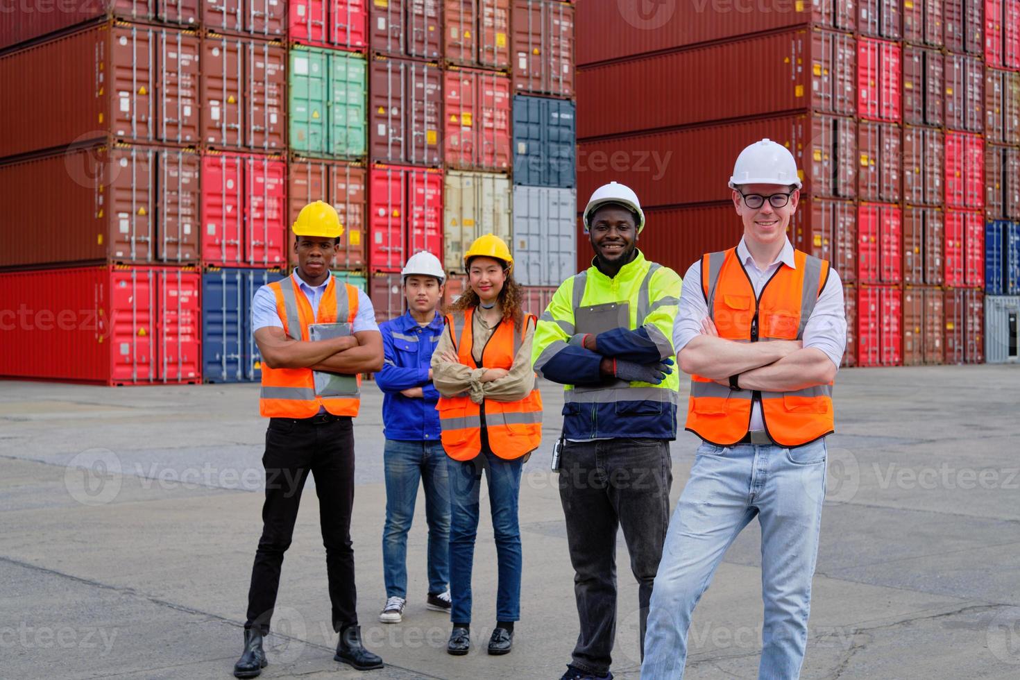 porträtt av en grupp multiracial arbetare team i säkerhetsuniformer, armarna i kors och tittar på kameran vid logistikdockan med många travar av containrar, fraktgods, godstransportindustri. foto