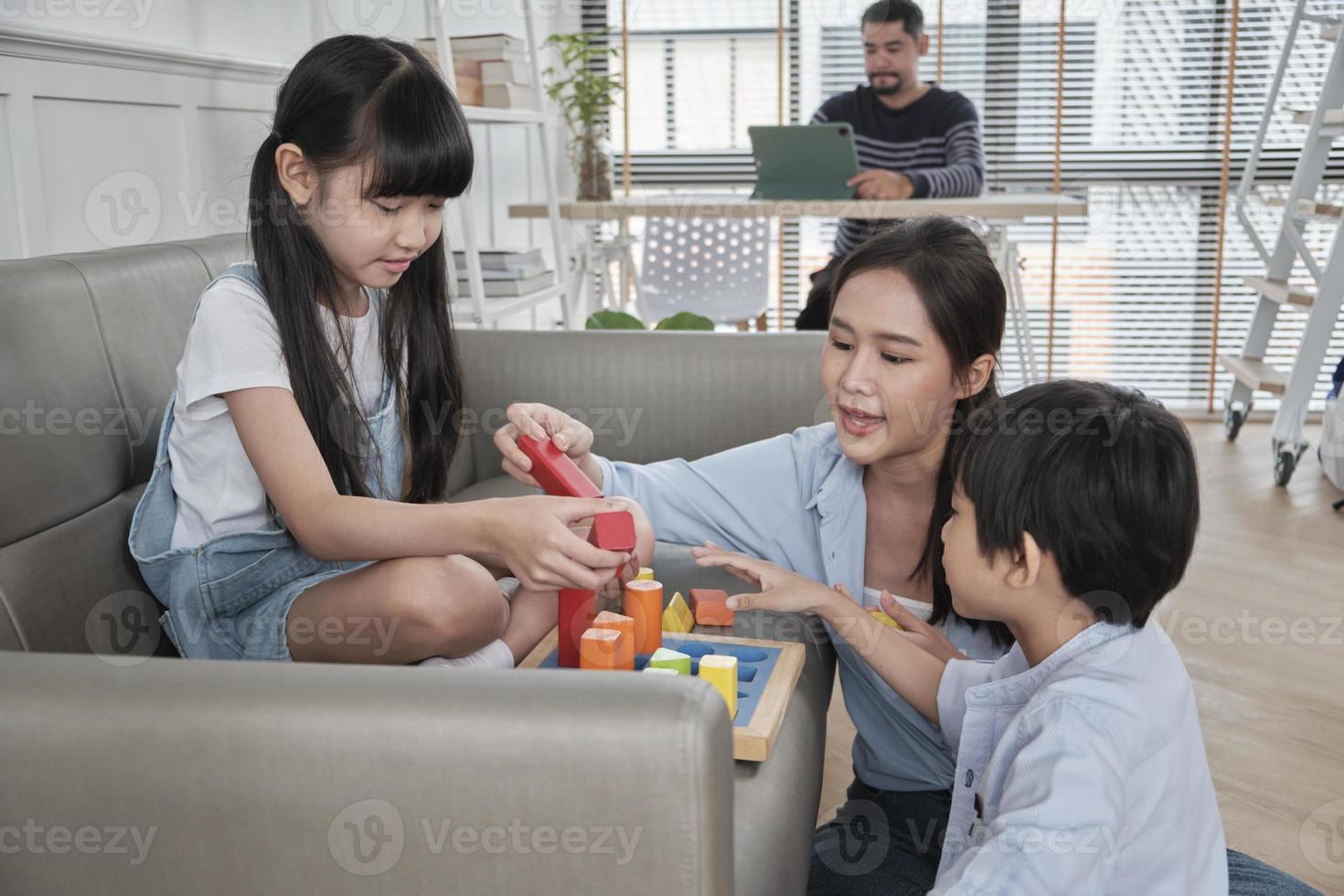 glad asiatisk thailändsk familjeomsorg, mamma och små barn har roligt när de leker med färgglada leksaksblock tillsammans på soffan i det vita vardagsrummet medan pappa jobbar, fritidshelg och välbefinnande livsstil. foto