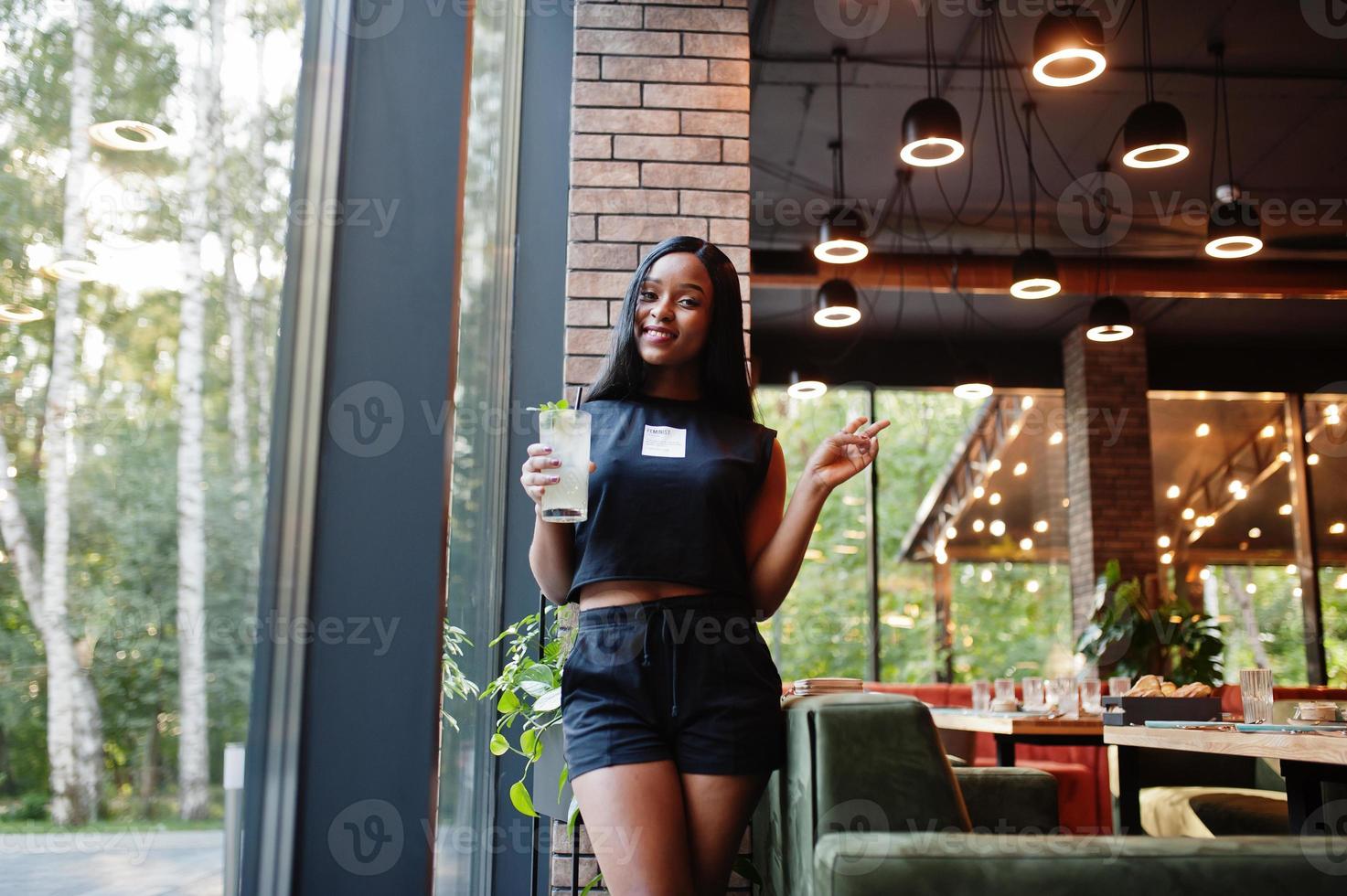 modern feminist afrikansk amerikan kvinna ha på sig i svart t-shirt och shorts, poserade på restaurang med citronsaft glas. foto