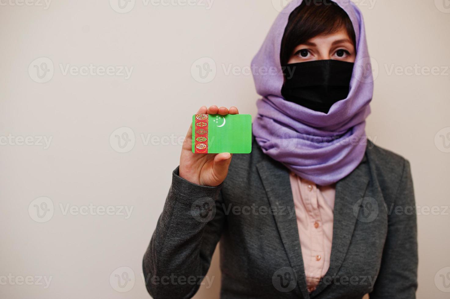 porträtt av ung muslim kvinna bär formell ha på sig, skydda ansikte mask och hijab huvud scarf, håll turkmenistan flagga kort mot isolerat bakgrund. coronavirus Land begrepp. foto