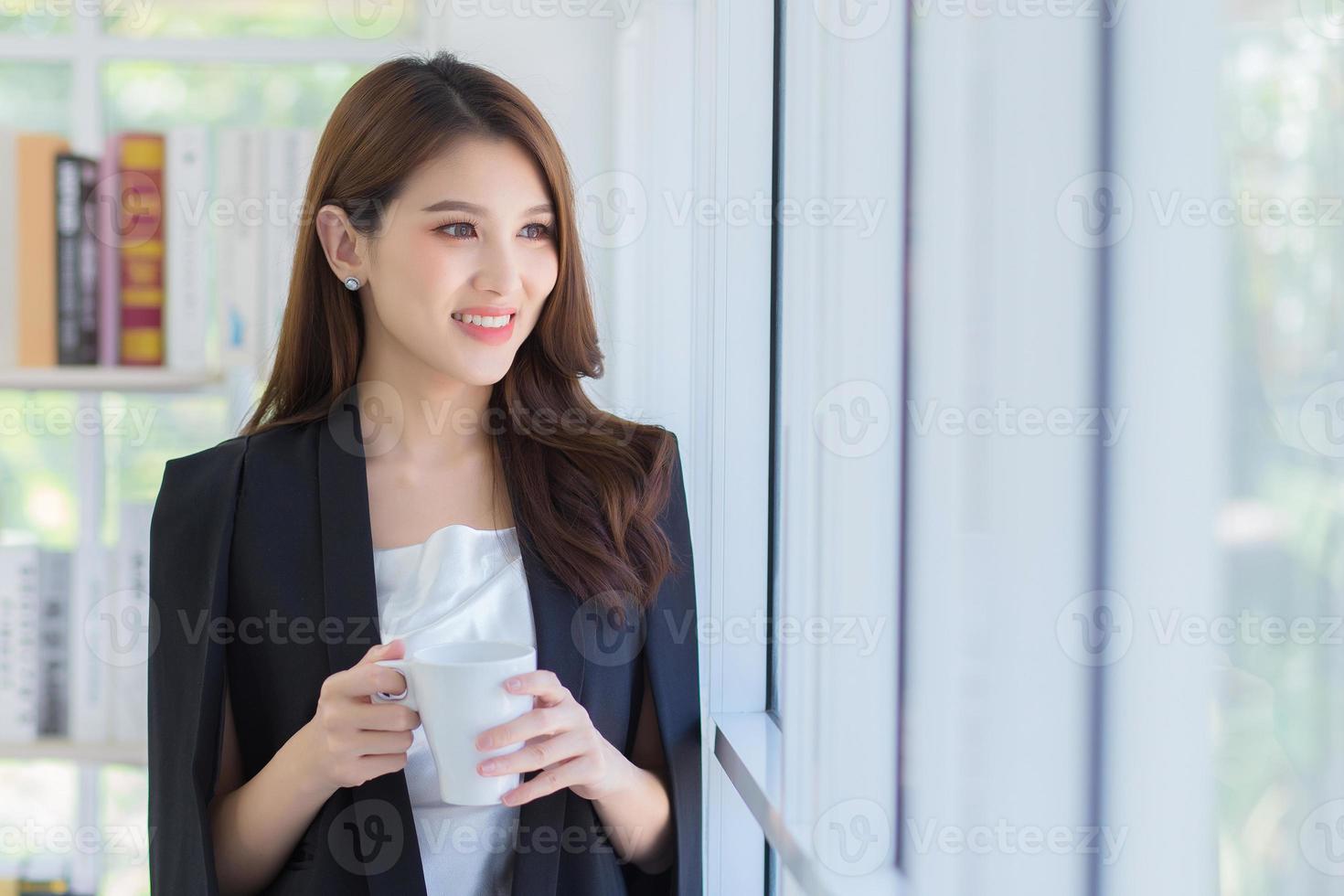 en skön kontor lady leende och ser ut av de fönster medan hon håll en kaffe kopp i henne händer. foto