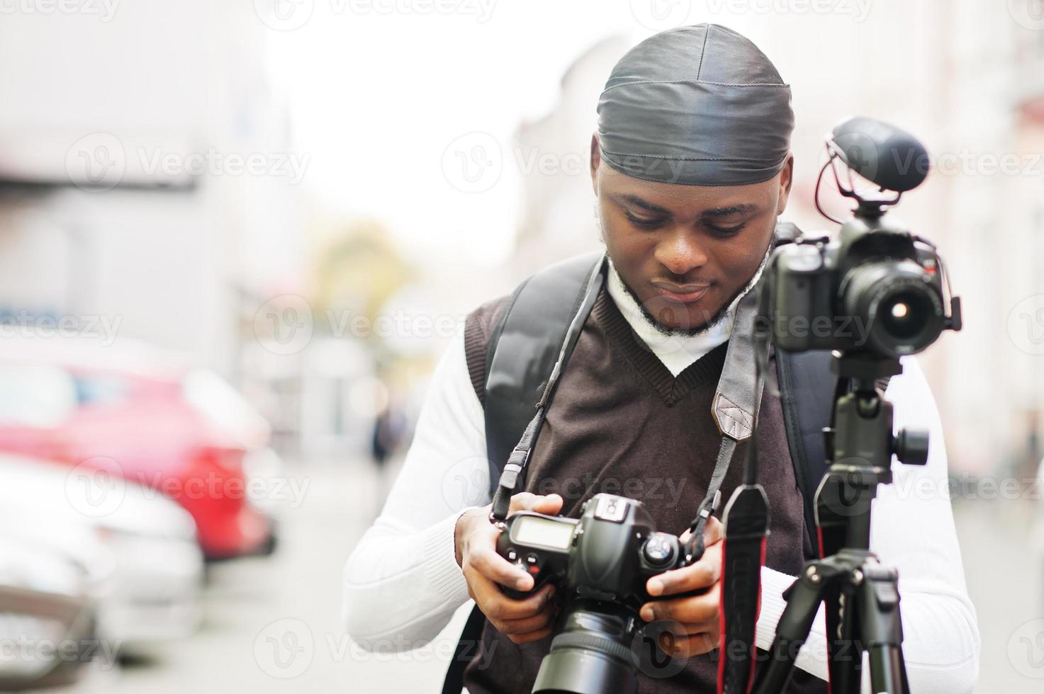 ung professionell afrikansk amerikan videographer innehav professionell kamera med stativ proffs Utrustning. afro kameraman bär svart duraq framställning en videoklipp. foto