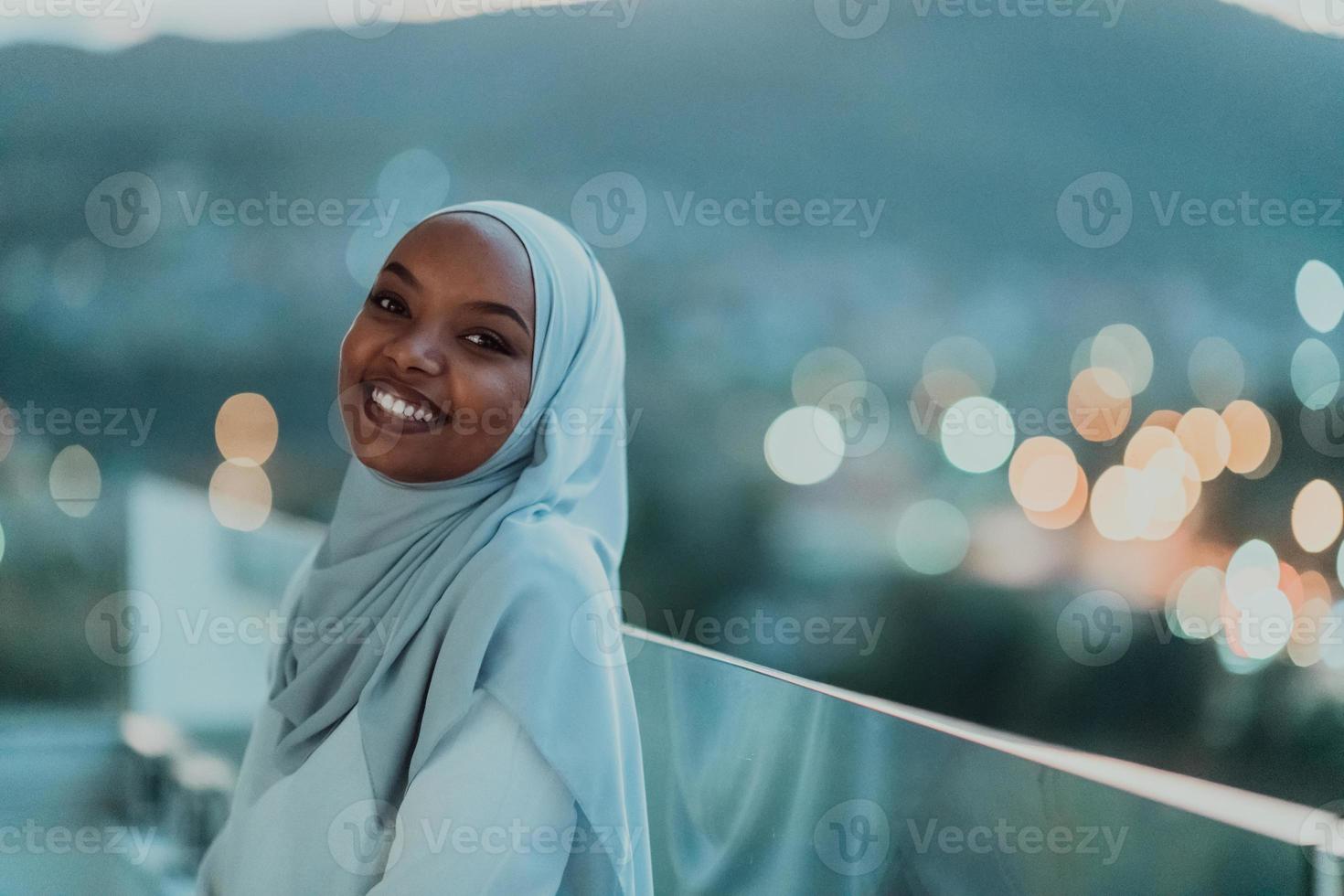 afrikansk muslim kvinna i de natt på en balkong leende på de kamera med stad bokeh lampor i de bakgrund. foto