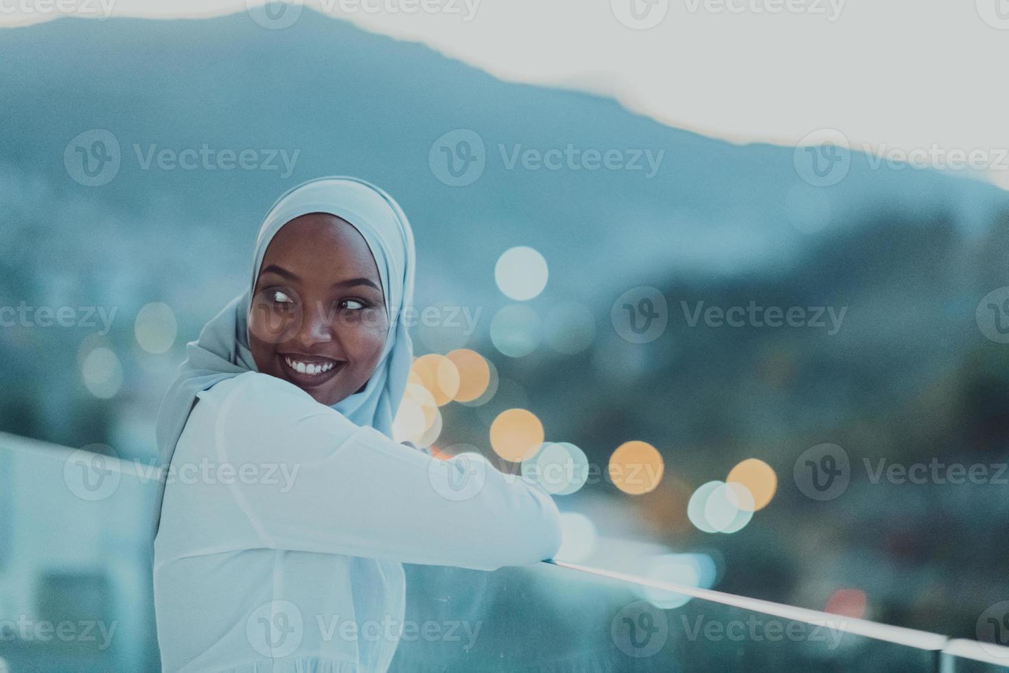 afrikansk muslim kvinna i de natt på en balkong leende på de kamera med stad bokeh lampor i de bakgrund. foto