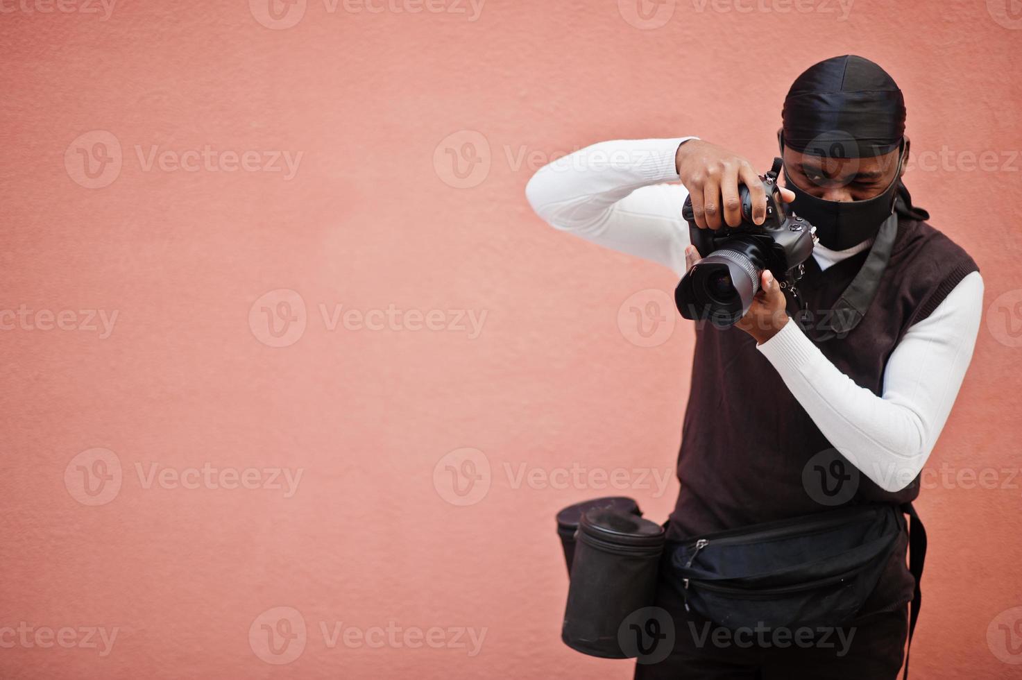 ung professionell afrikansk amerikan videographer innehav professionell kamera med proffs Utrustning. afro kameraman bär svart duraq och ansikte skydda mask, framställning en videoklipp. foto