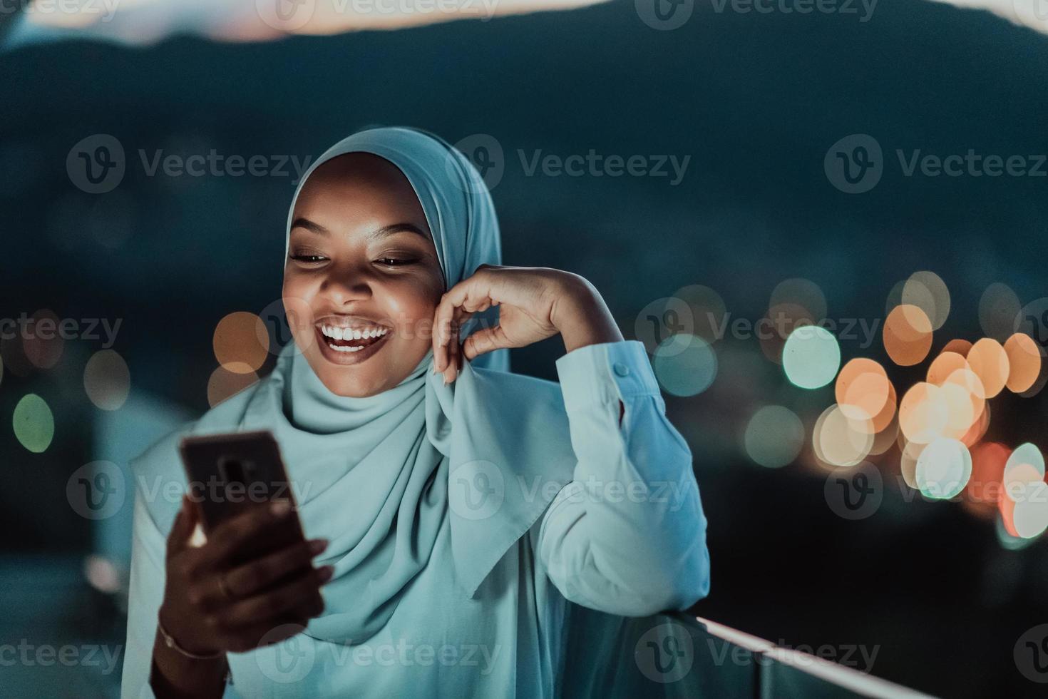 ung muslim kvinna bär scarf slöja på urban stad gata på natt textning på en smartphone med bokeh stad ljus i de bakgrund. foto