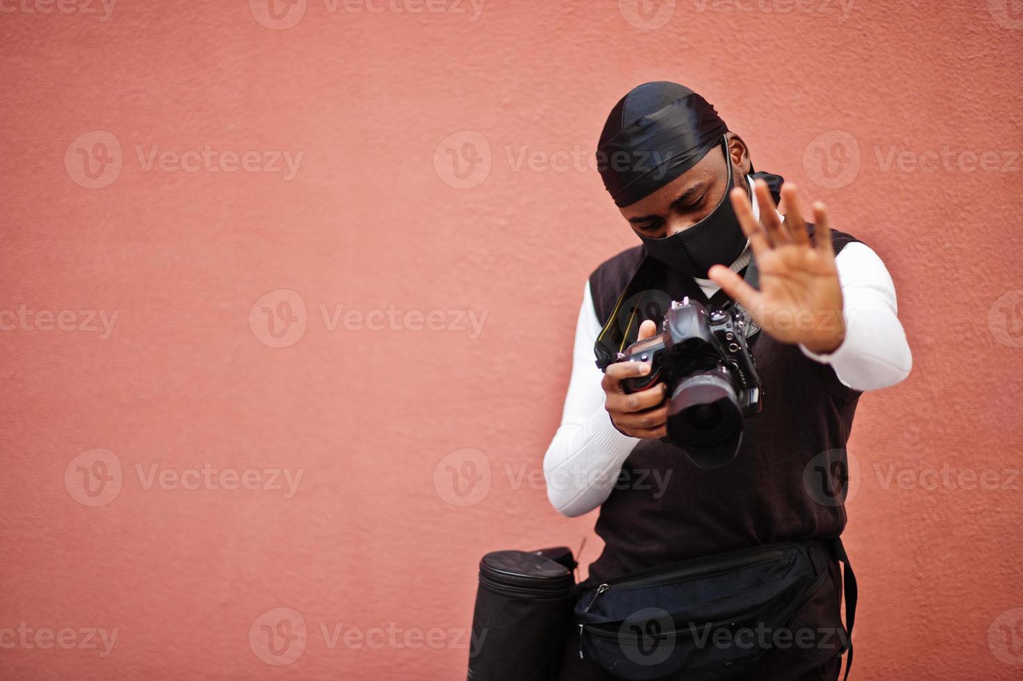 ung professionell afrikansk amerikan videographer innehav professionell kamera med proffs Utrustning. afro kameraman bär svart duraq och ansikte skydda mask, framställning en videoklipp. foto