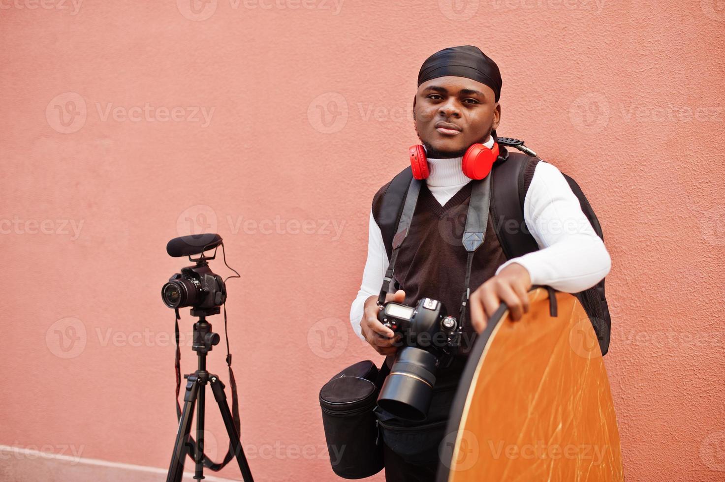 ung professionell afrikansk amerikan videographer innehav professionell kamera med proffs Utrustning. afro kameraman bär svart duraq framställning en videoklipp. foto
