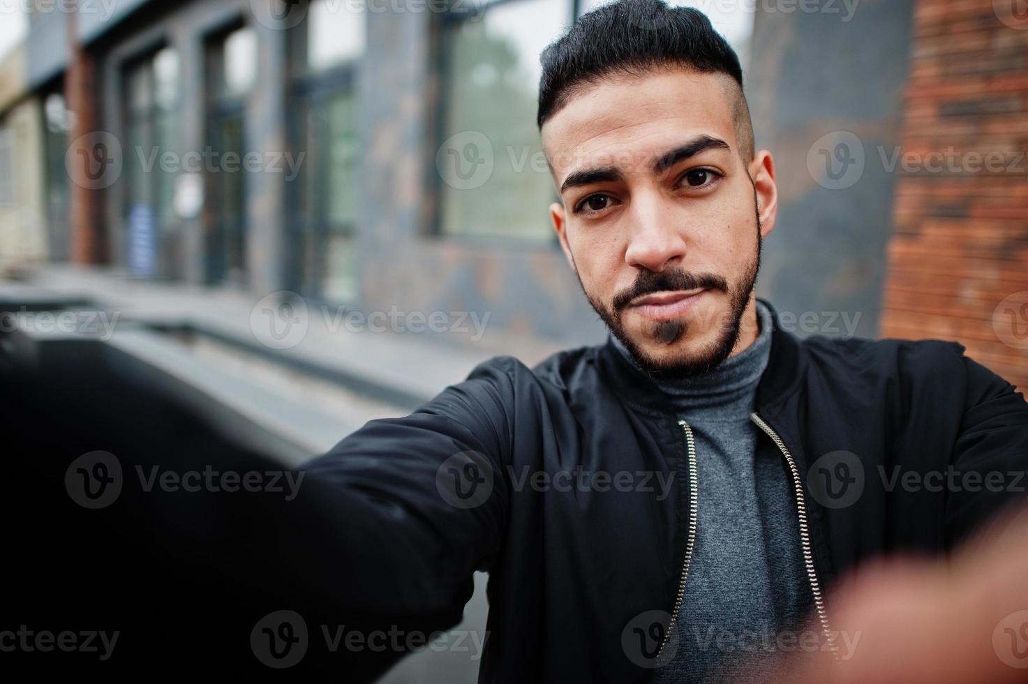 porträtt av eleganta arab skägg man ha på sig grå turtleneck och svart jacka. arab modell kille göra selfie förbi kamera. foto