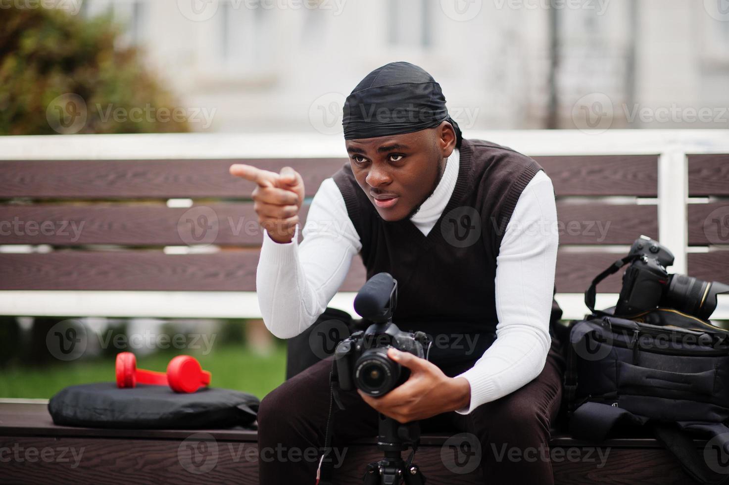 ung professionell afrikansk amerikan videographer innehav professionell kamera med proffs Utrustning. afro kameraman bär svart duraq framställning en videoklipp. foto