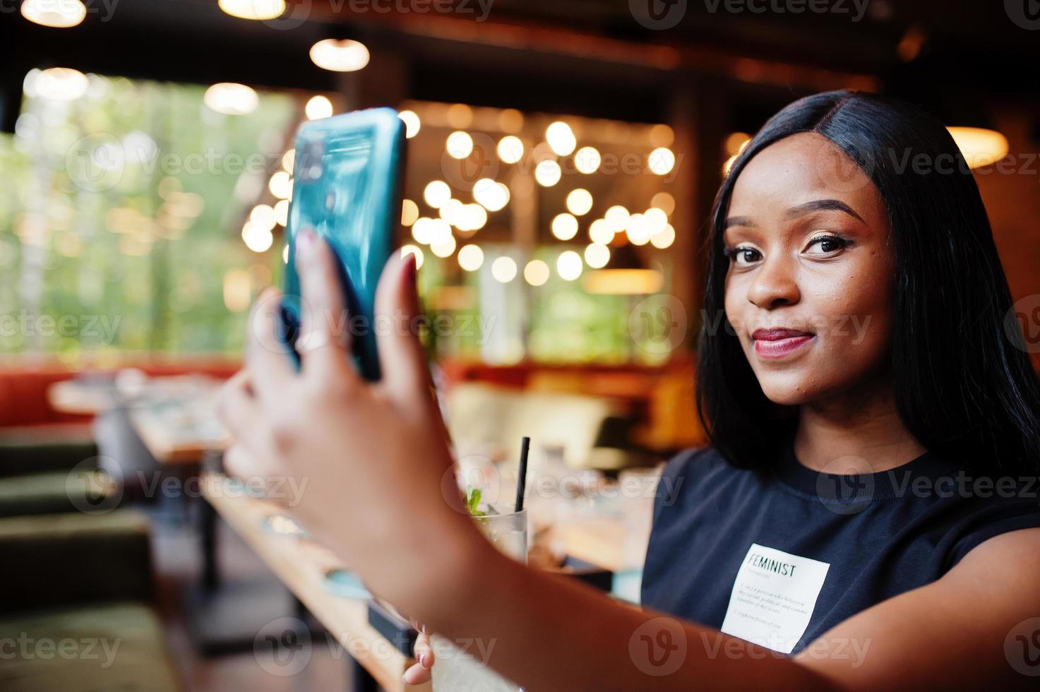 modern feminist afrikansk amerikan kvinna ha på sig i svart t-shirt och shorts, framställning selfie på restaurang med citronsaft glas och mobil telefon. foto