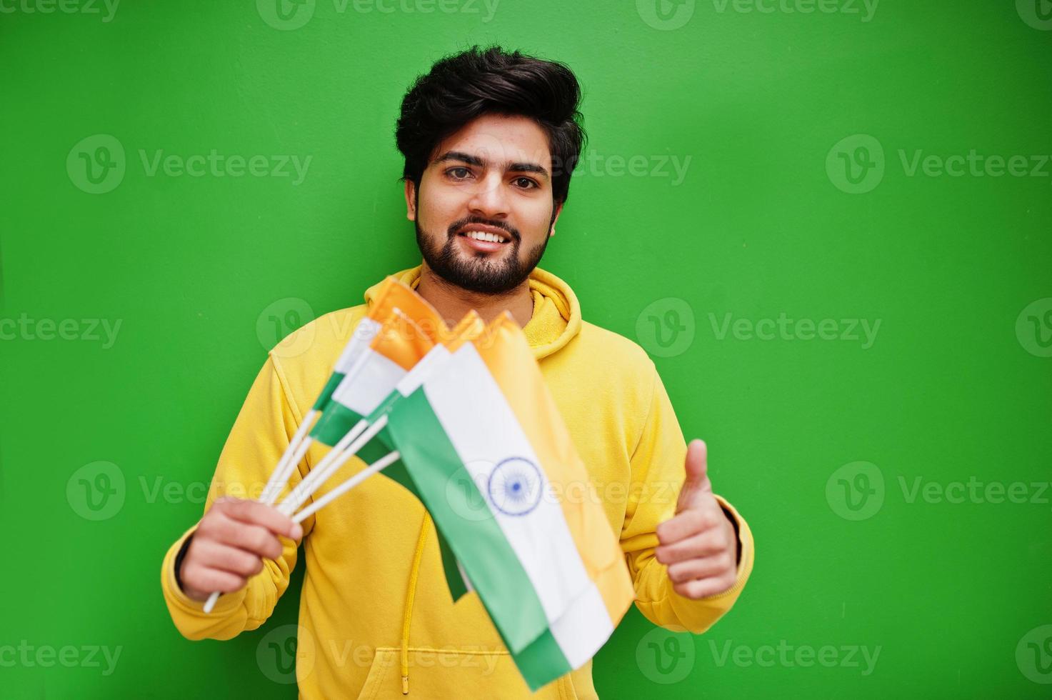 urban ung hipster indisk man i en modern gul tröja håll flaggor av Indien i händer. Häftigt söder asiatisk kille ha på sig luvtröja isolerat på grön bakgrund. foto