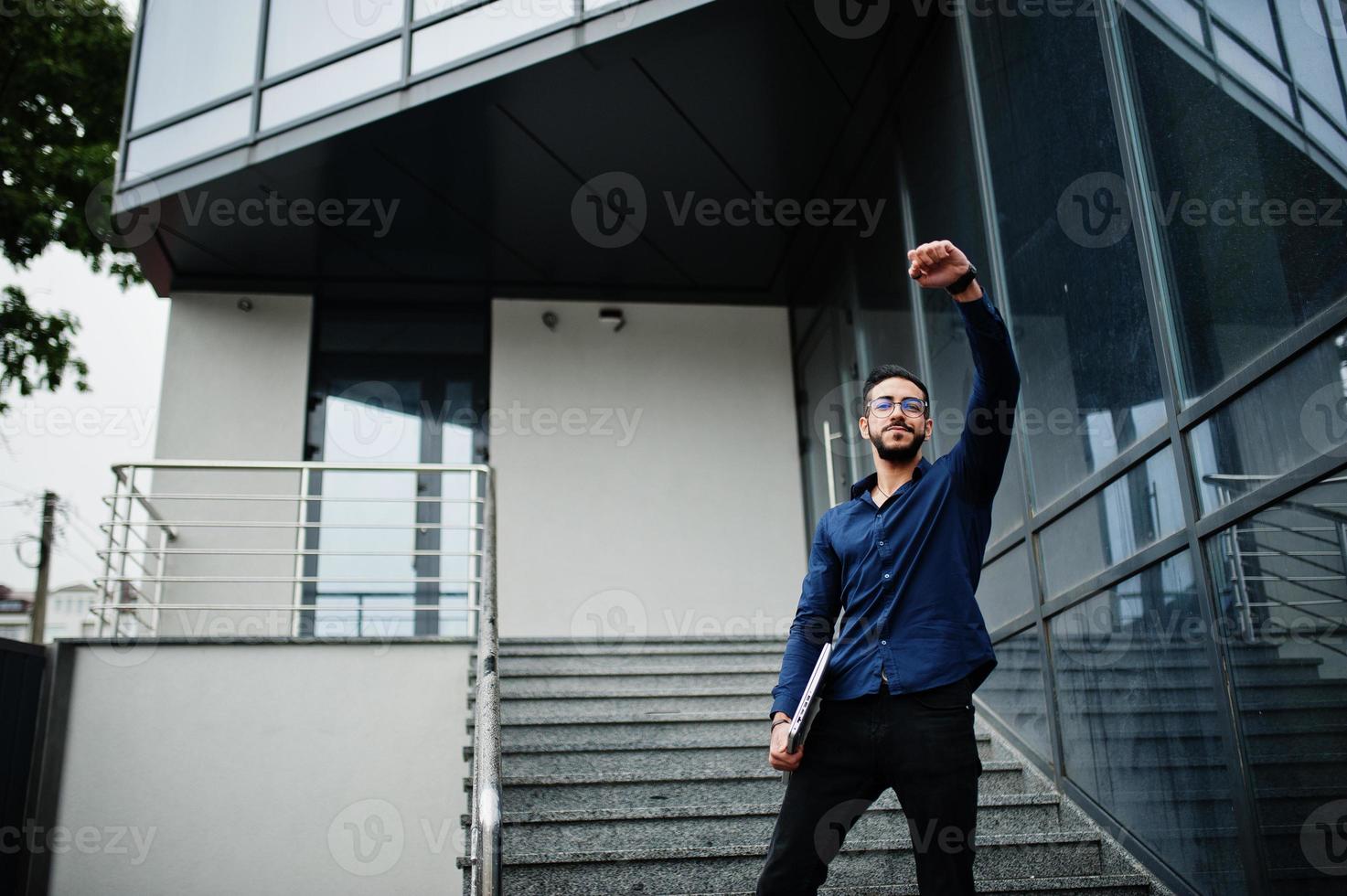 mitten östra entreprenör ha på sig blå skjorta, glasögon med bärbar dator mot kontor byggnad. foto