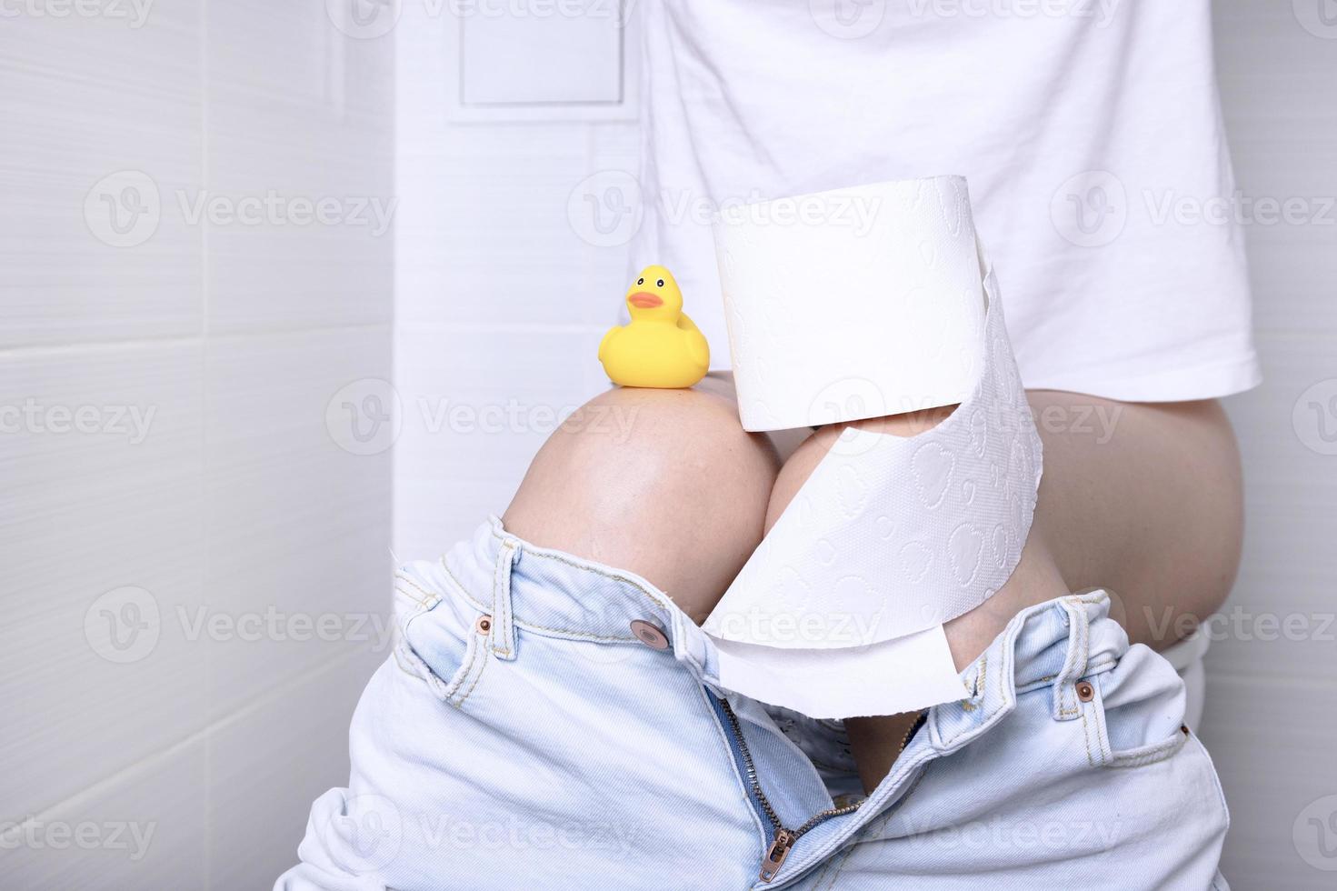 pissande kvinna lidande från diarre i en vit toalett, i en toalett med en rulla av vit papper med en gul Anka i henne hand. foto