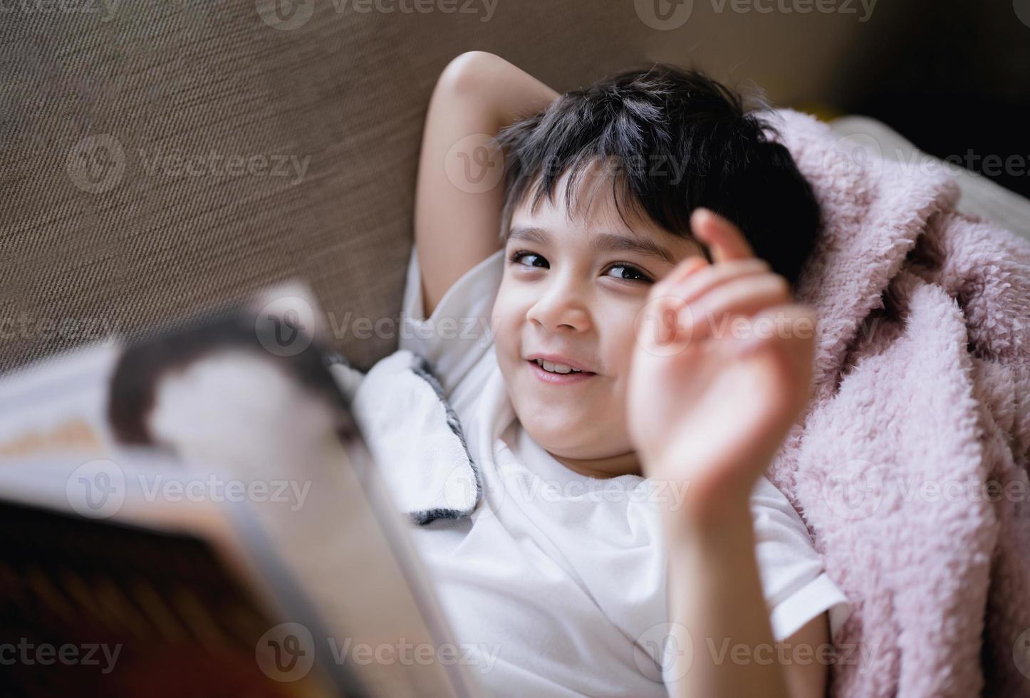 Lycklig skola unge liggande på soffa som visar hand och ser upp med leende ansikte, barn pojke läsning berättelse avkopplande på Hem på helgen, positiv barn begrepp foto