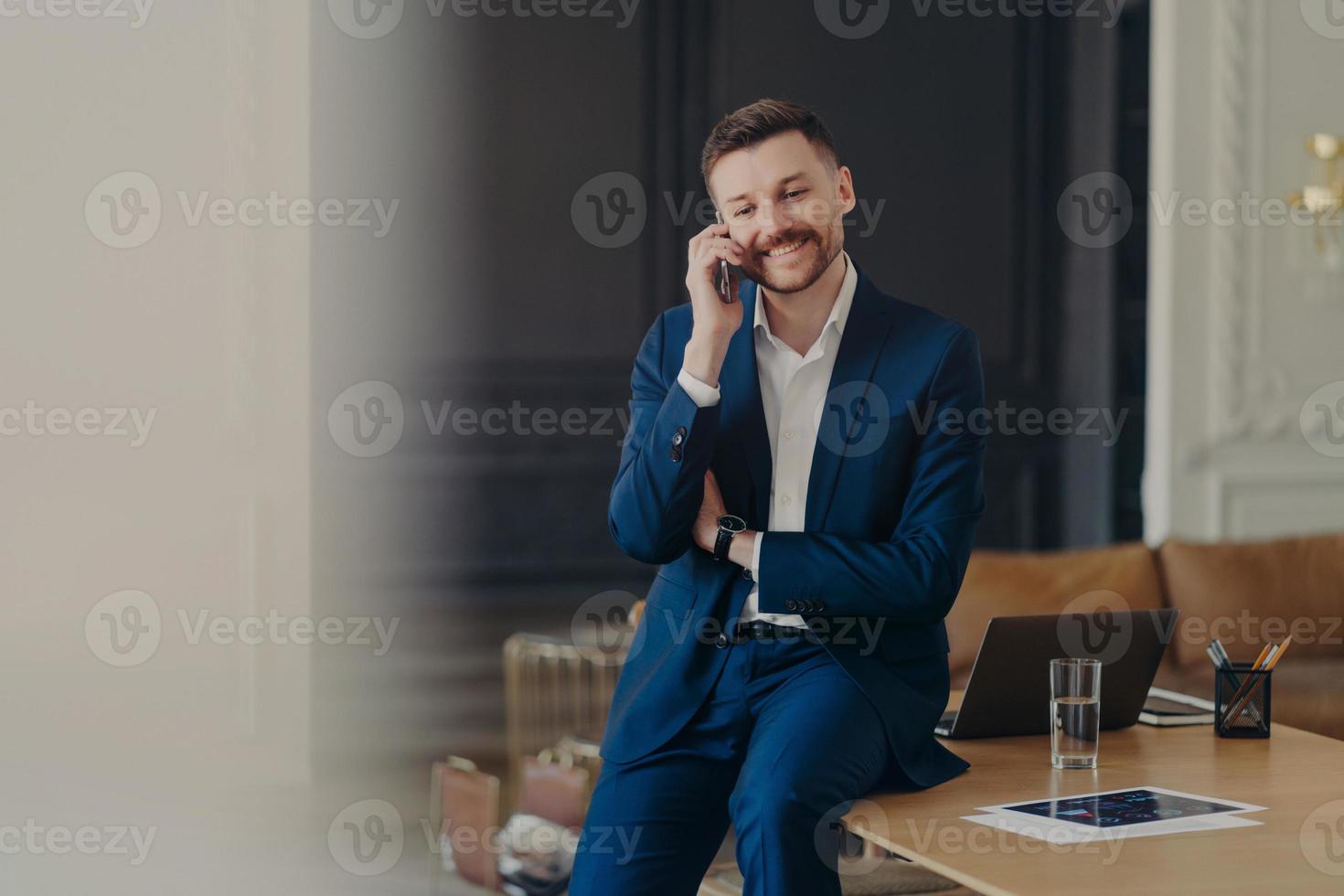 glad stilig affärsman pratar i telefon när han sitter på skrivbordet foto