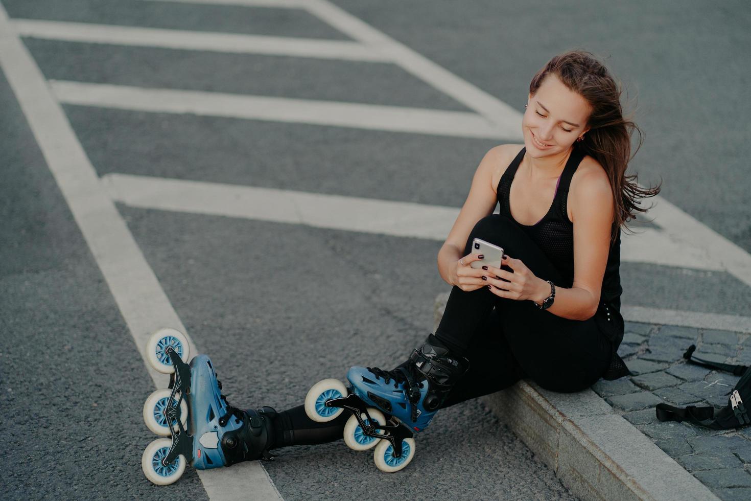 ovanifrån av brunett aktiv ung kvinna använder smartphone för onlinekommunikation poserar på asfalt klädd i svart aktivt slitage åtnjuter favorithobby och fritid. extrem sport och vila koncept foto