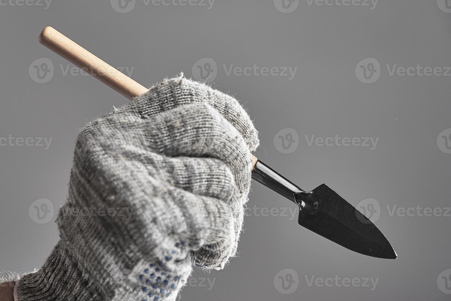 hand i handske innehav trädgårdsarbete spade tycka om en penna på grå bakgrund. foto