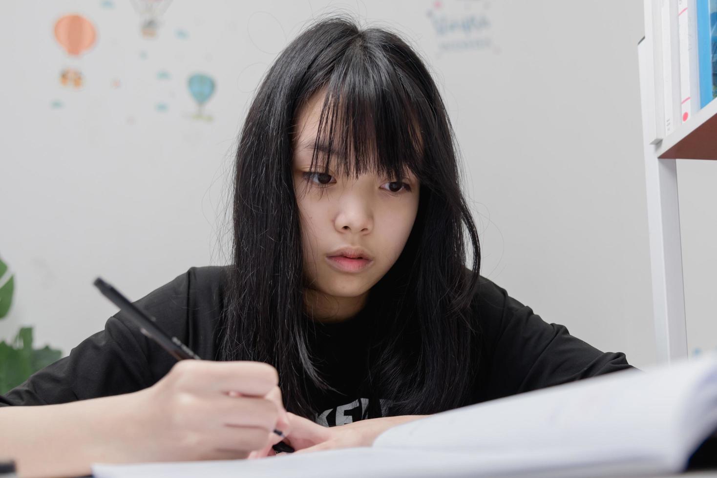 asiatisk studentflicka skriver läxor och läser bok vid skrivbordet foto