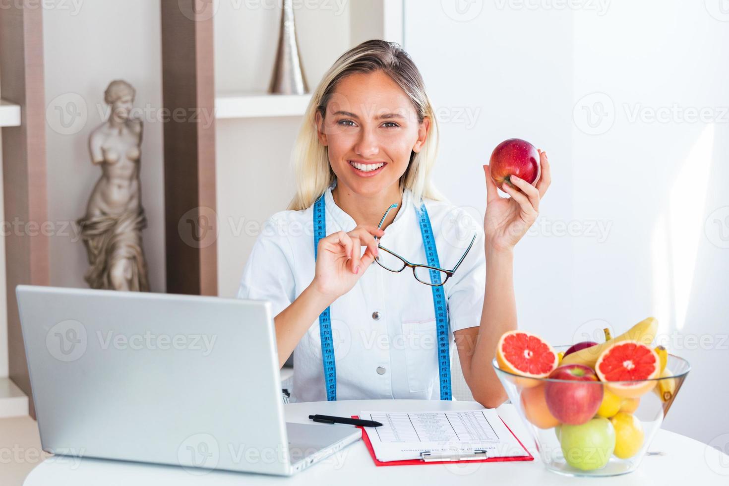 porträtt av ung leende kvinna dietist i de samråd rum. dietist skrivbord med friska frukt, juice och mätning tejp. dietist arbetssätt på diet planen. foto