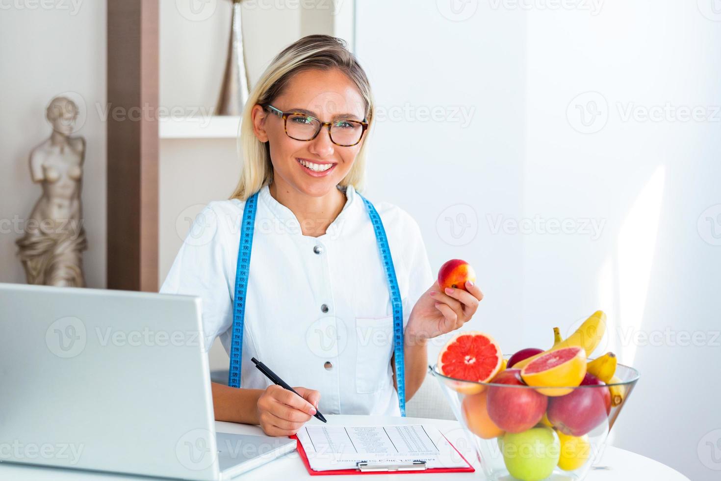leende dietist i henne kontor, hon är som visar friska grönsaker och frukter, sjukvård och diet begrepp. kvinna dietist med frukt arbetssätt på henne skrivbord. foto
