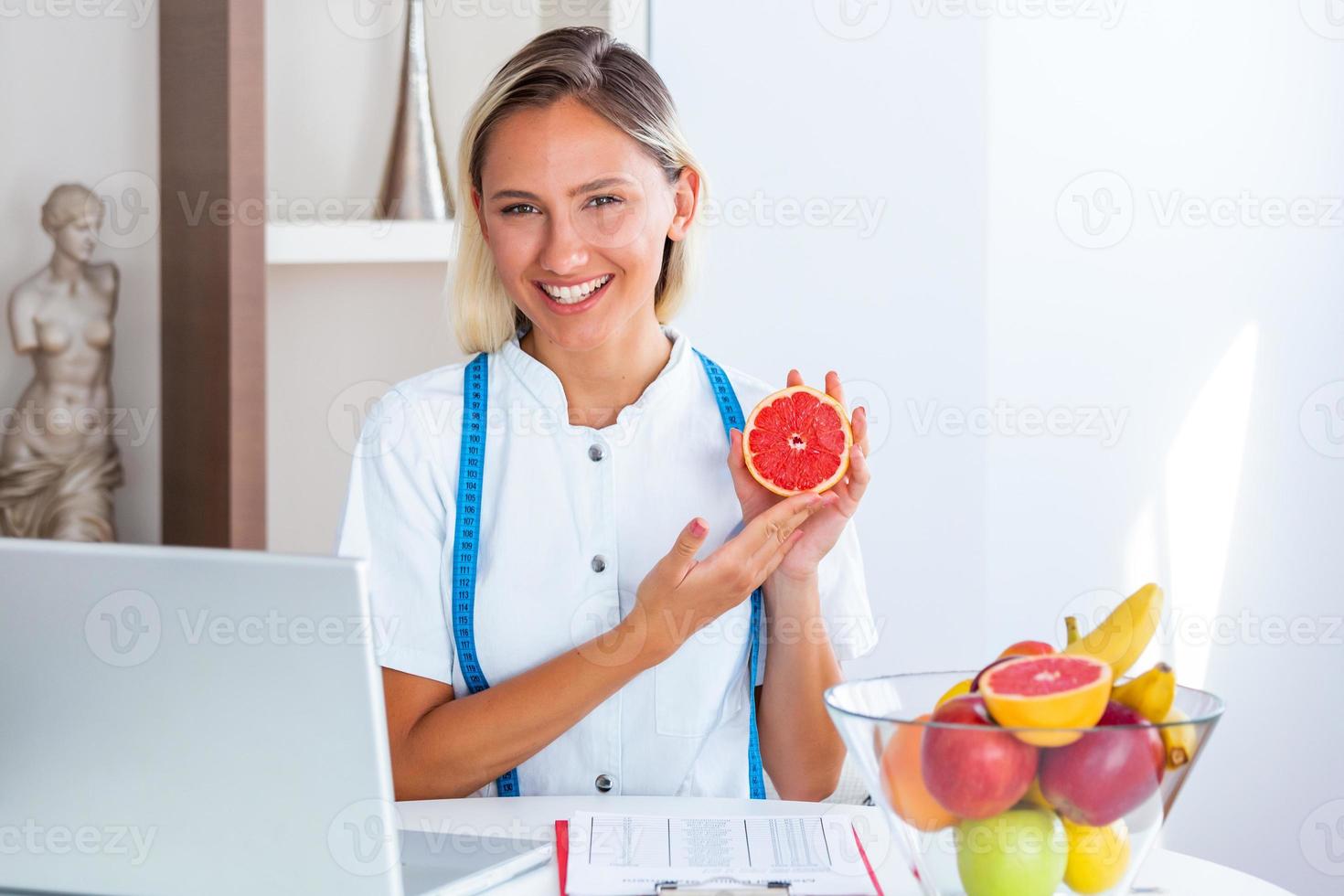 porträtt av ung leende kvinna dietist i de samråd rum. dietist skrivbord med friska frukt, juice och mätning tejp. dietist arbetssätt på diet planen. foto