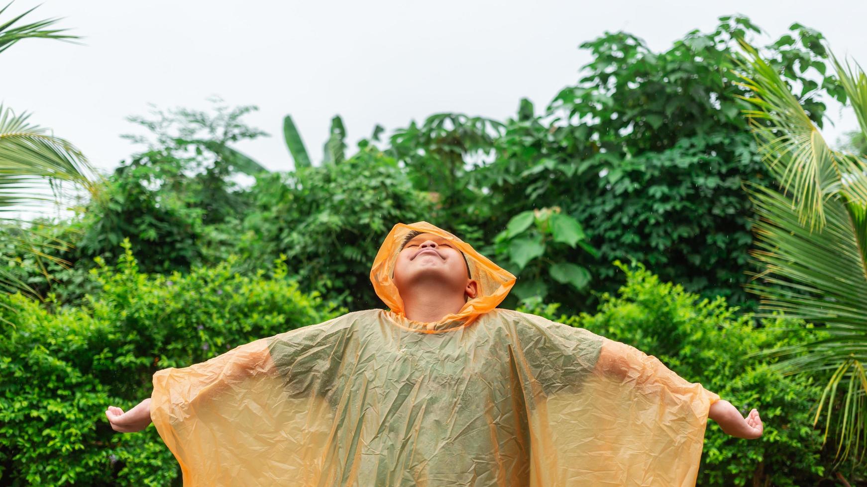 asiatisk pojke bär orange regnkappa är Lycklig och har roligt i de regn på en regnig dag. foto