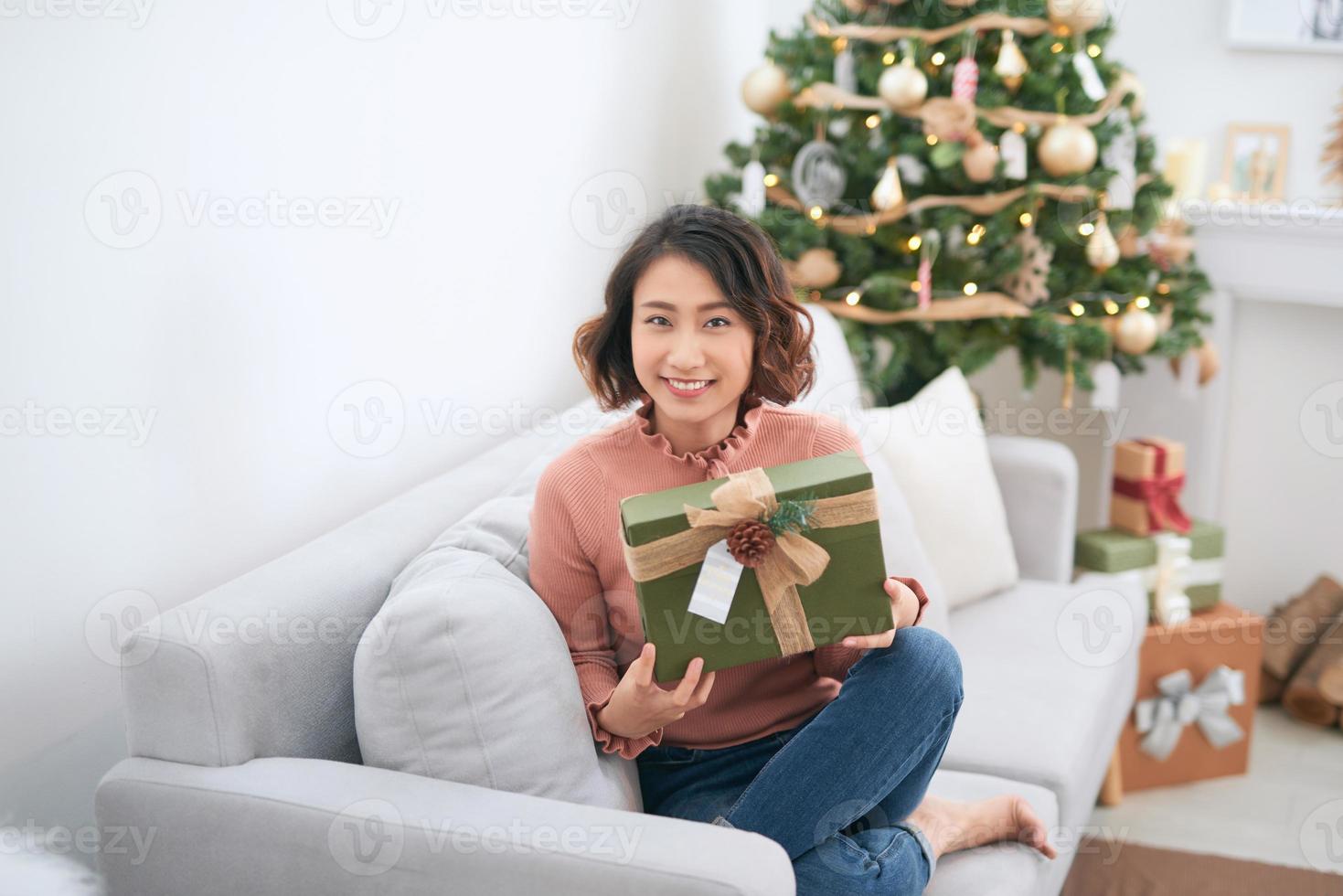 skön flicka innehav en jul närvarande. Lycklig kvinna stående nära ny år träd och väntar för firande. foto