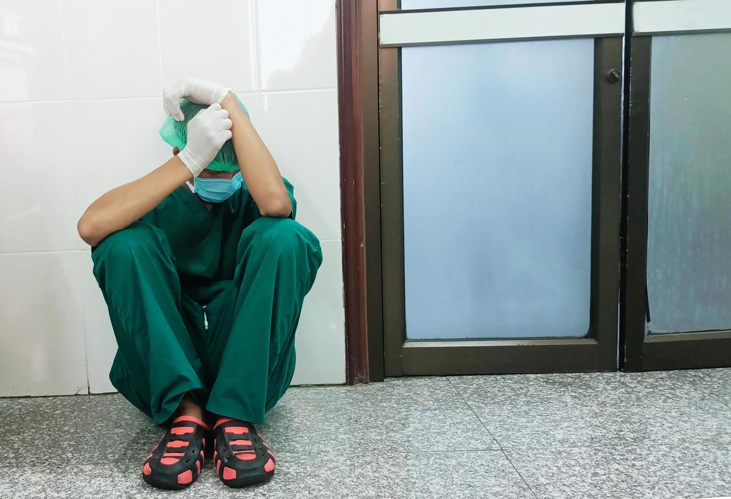 en trött utmattad manlig läkare bär en ansikte mask och blå enhetlig handske sitta på de sjukhus golv. de sänka ledsen svart etik- läkare känna Trötthet burnout påfrestning, brist av sova, och tupplur på arbete. foto