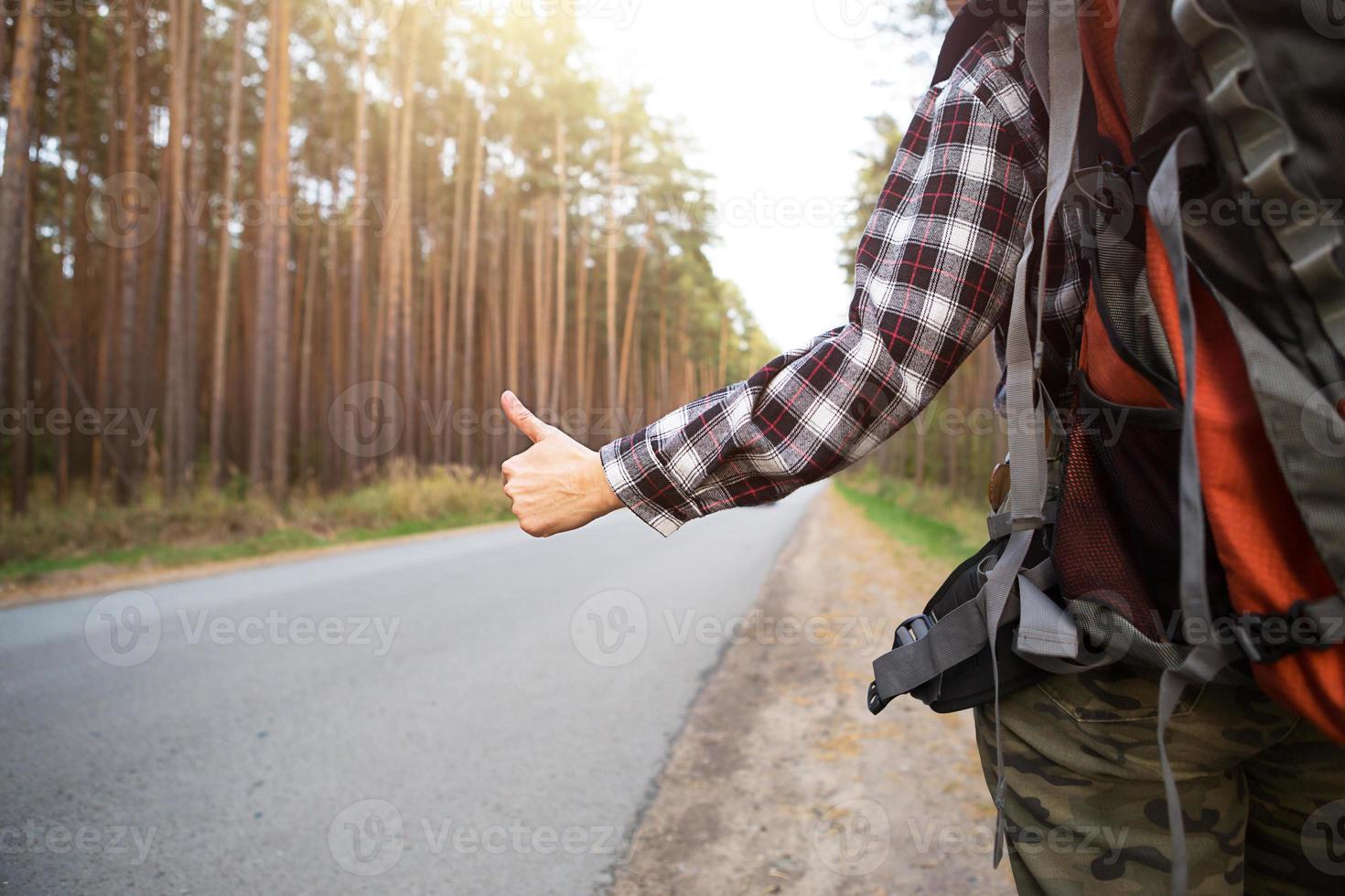 hand med tummen upp på bakgrunden av motorvägen - lifta, rösta. turist i en rutig skjorta med ryggsäck nära skogen lifta en tur. lifta, inhemsk turism, ensam äventyr, resa, vandring foto