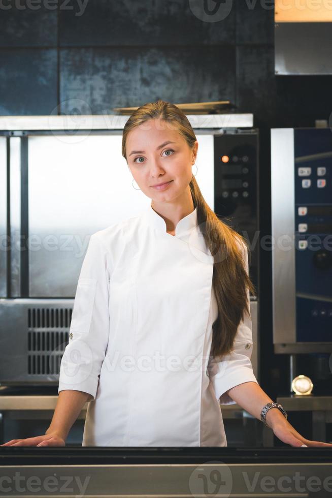 porträtt av självsäker och leende ung kvinna kock klädd i vit enhetlig, professionell kök är på bakgrund foto