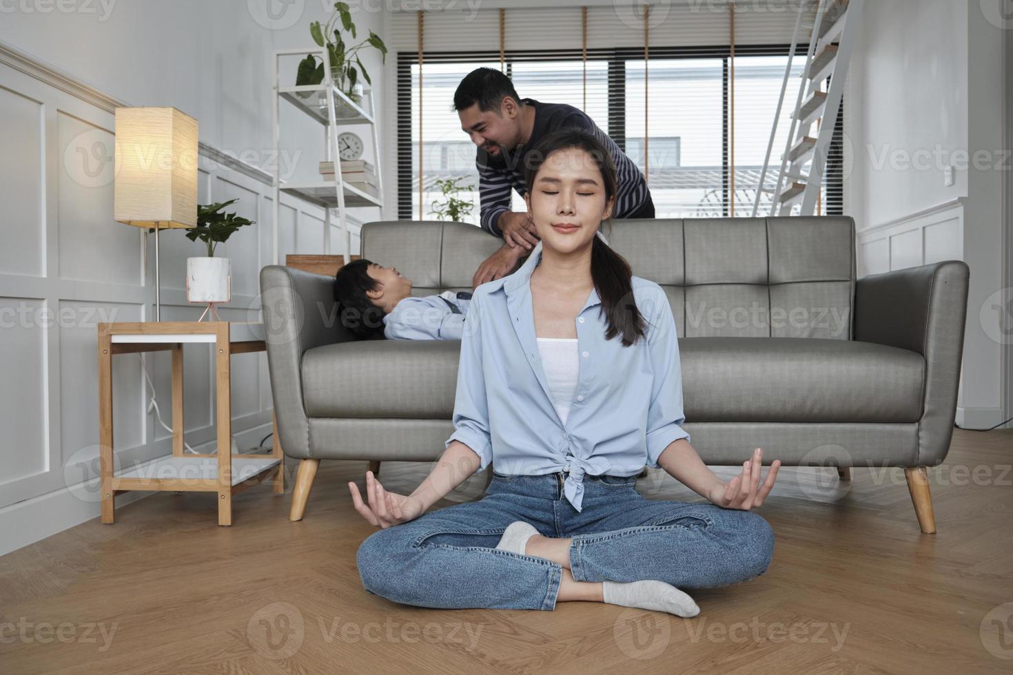 ung asiatisk thai mor sitter på levande rum golv, mediterar, och praxis yoga för hälsa och wellness när Make och son reta tillsammans på soffa, Lycklig inhemsk Hem livsstil på familj helgen. foto