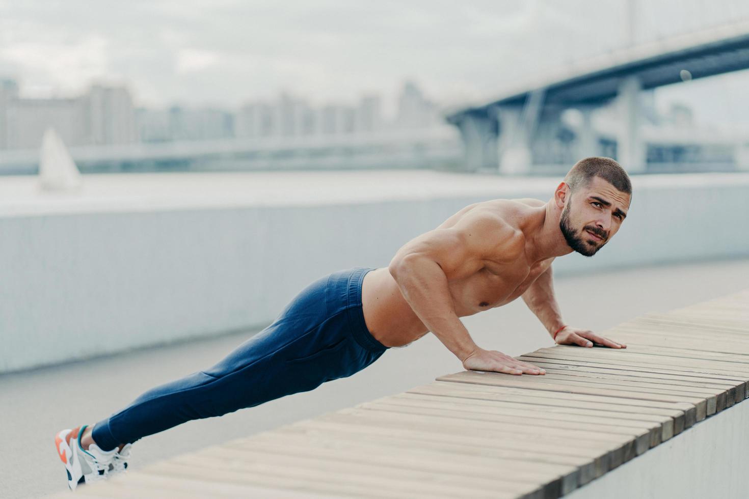 full längd bild av sportig muskulös man i sportkläder gör push up träning har koncentrerat allvarliga uttryck poser utomhus. självbestämd skäggig idrottsman planking för att ha starka armar foto