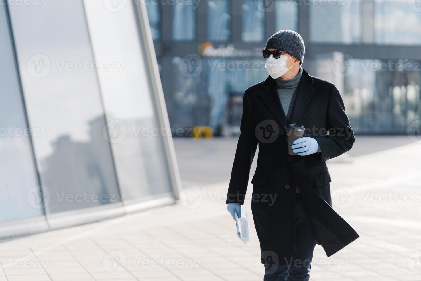 horisontell bild av allvarlig vuxen man som bär engångskopp kaffe, klädd i ytterkläder, skyddande medicinsk mask och handskar, skyddar sig själv under coronavirus-pandemin, koncentrerad åt sidan foto