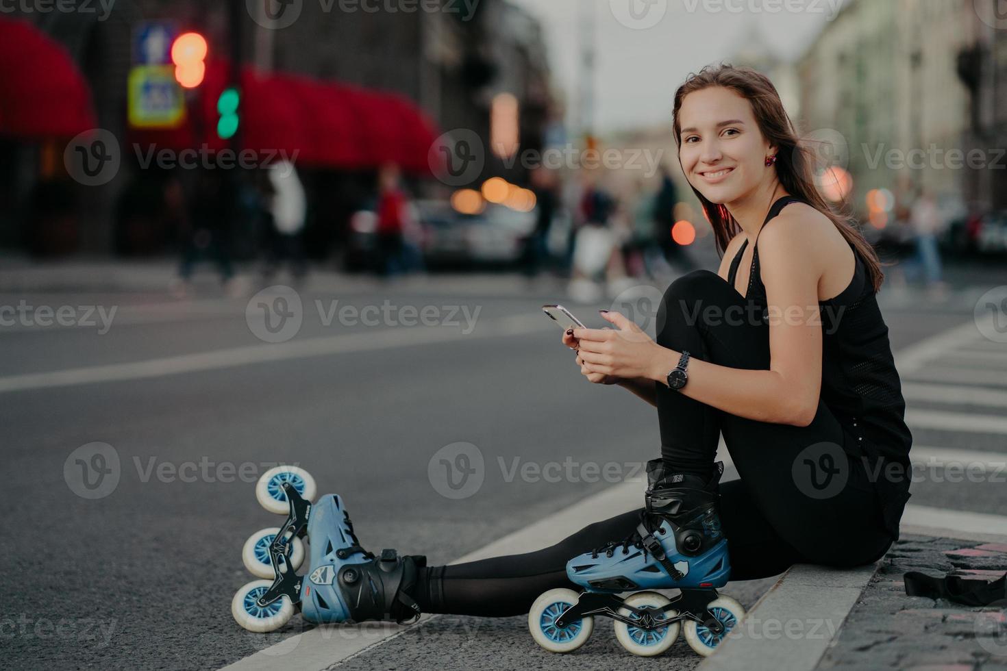 människor utomhus- aktiviteter och rekreation begrepp. horisontell skott av aktiva smal kvinna varelse i Bra fysisk form rider rullskridskor användningar smartphone skickar text meddelanden uppkopplad poser utanför foto