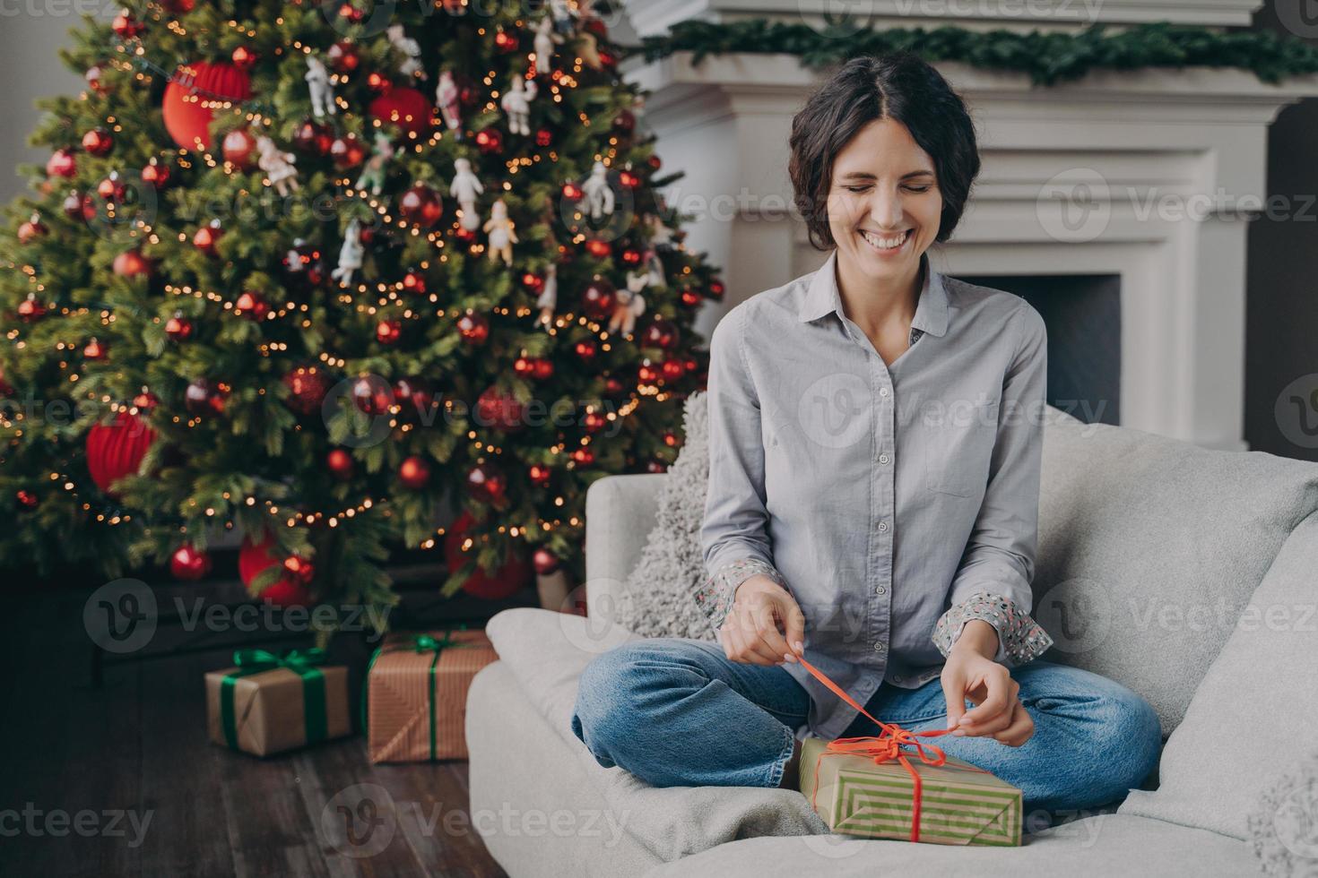 upprymd italiensk kvinna med slutna ögon som försöker packa upp julklappen när hon sitter på soffan hemma foto