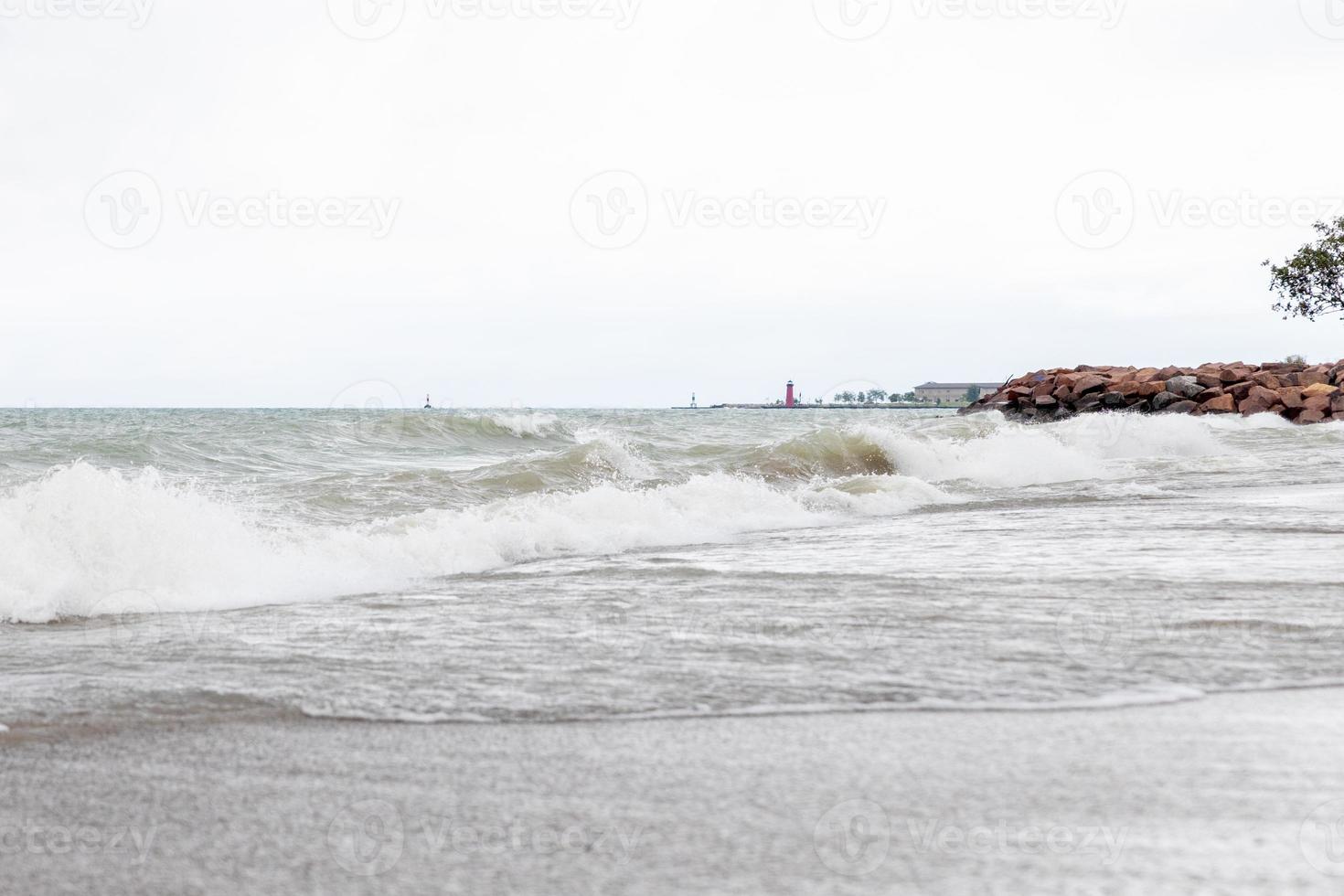 stor vågor rullande in i de sandig strandlinje av sjö Michigan på en molnig mulen dag. stenar innehav de strandlinje tillbaka med träd i de hörn. byggnader och fyr sett i de distans. foto