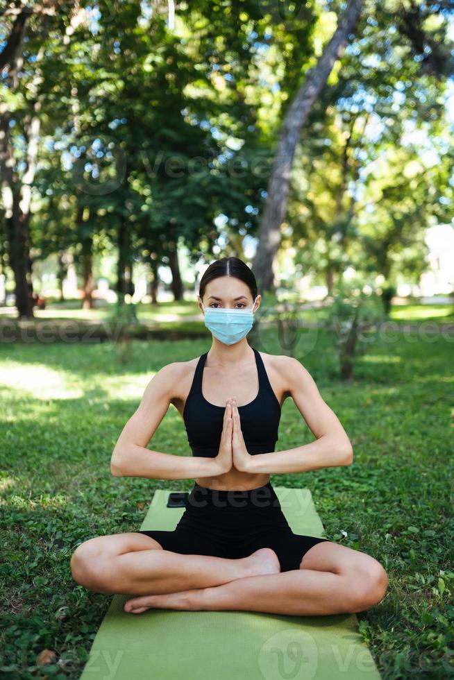 atletisk ung kvinna i en medicinsk skyddande mask, håller på med yoga i de parkera foto