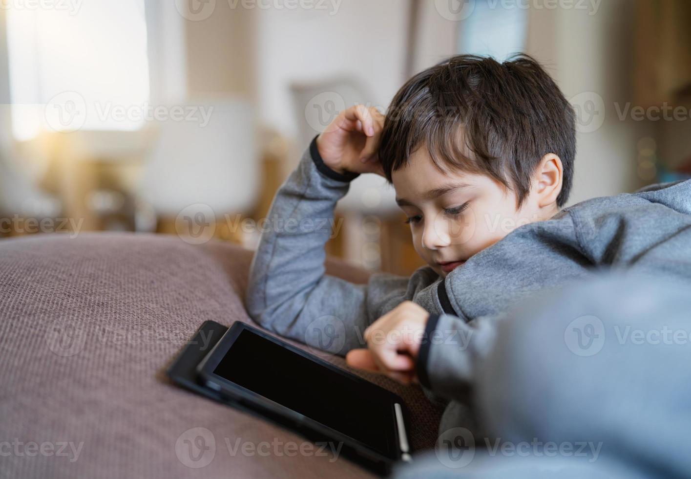 äkta unge Sammanträde på soffa tittar på tecknade serier eller spelar spel på surfplatta, barn pojke använder sig av digital vaddera inlärning lektion på internet, hem skolgång, distans inlärning uppkopplad utbildning begrepp foto