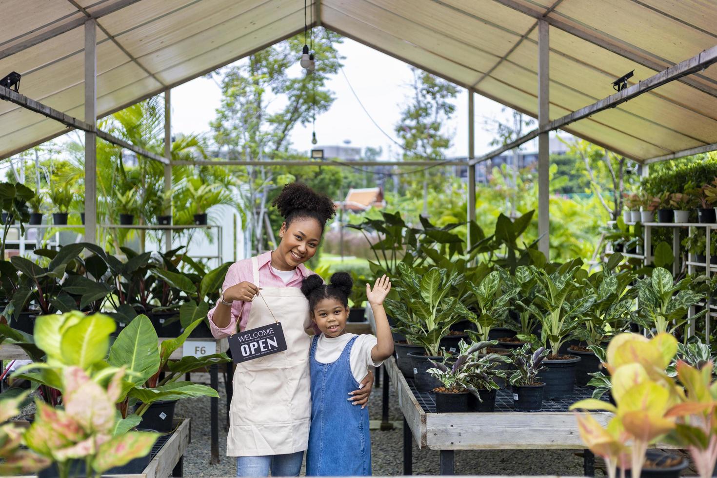 afrikansk trädgårdsmästare och ägare med henne dotter är innehav öppen tecken i främre av deras dekorativ krukväxt på deras egen barnkammare trädgård Centrum full av exotisk och tropisk växt begrepp foto