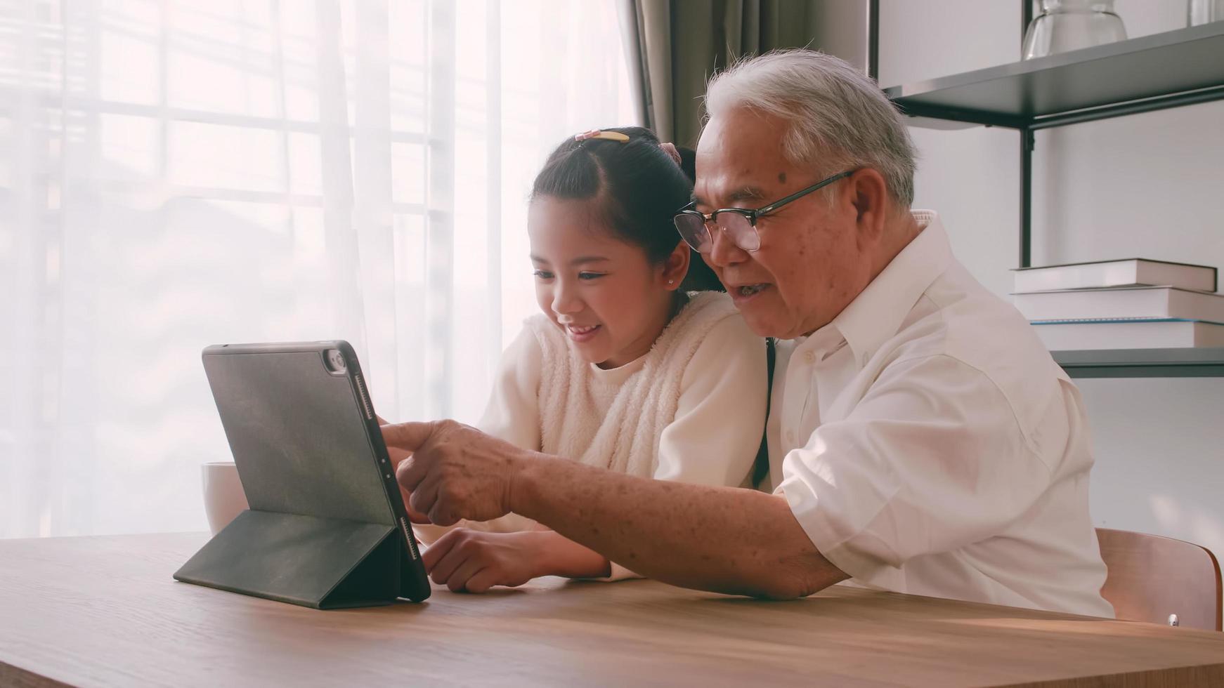 ett asiatisk farfar och barnbarn utgifterna tid tillsammans i de levande rum. Lycklig senior man med liten flicka använder sig av en läsplatta på de tabell på Hem. foto