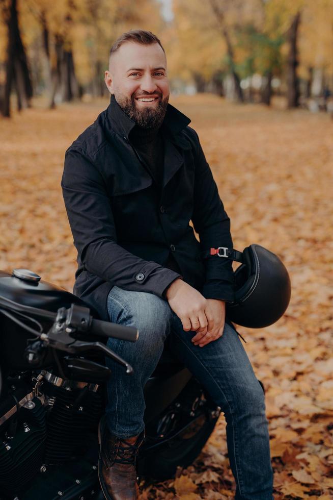 vertikal bild av en leende motorcyklist ler glatt, håller i hjälmen, poserar på motorcykeln, bär svart jacka och jeans, kör genom parken, njuter av hösten och åker svart fordon. transport koncept foto