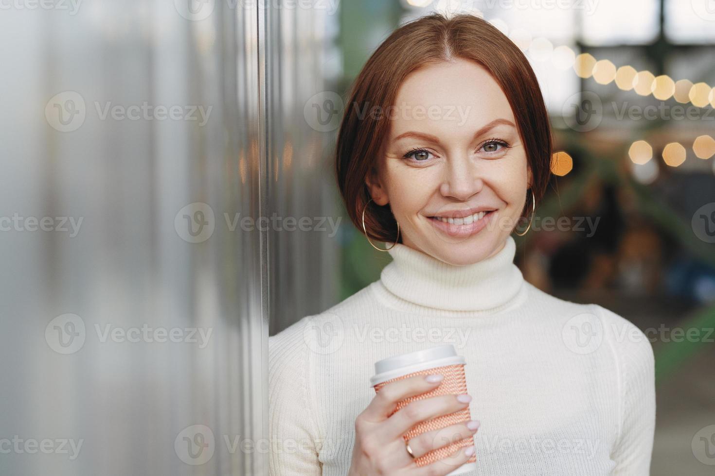 snygg ung kvinna klädd i avslappnad vit polotröja, håller en papperskopp med aromatisk cappucino eller kaffe, tittar glatt på kameran, poserar utanför, har fritid. livsstilskoncept foto