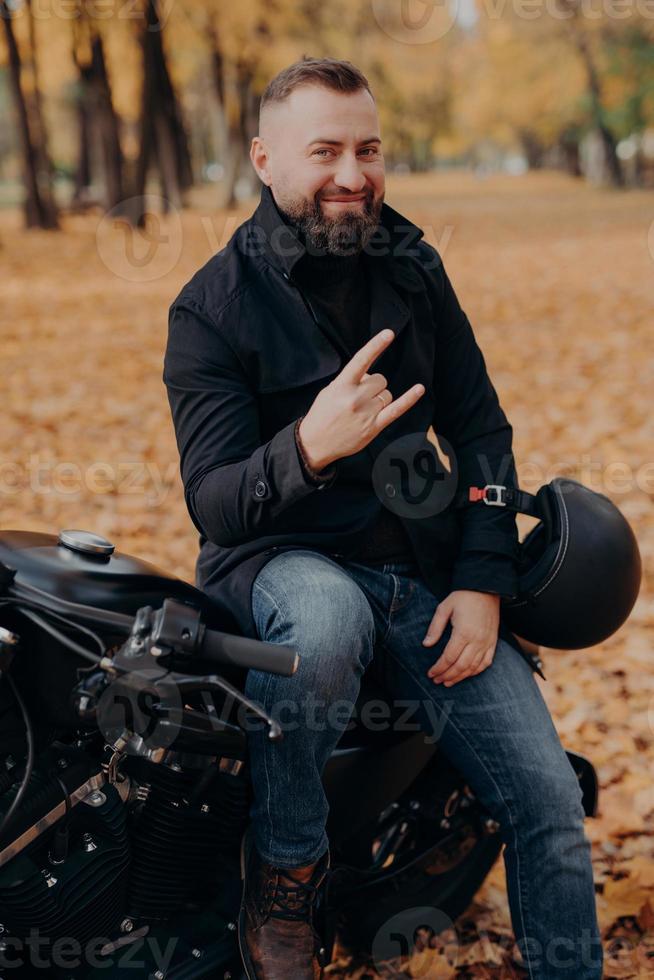 stilig skäggig manlig motorcyklist håller hjälm, gör horngest med fingrar, känns cool, bär svart kappa och jeans, sitter på snabb motorcykel mot höstens parkbakgrund. bekymmerslös racer foto