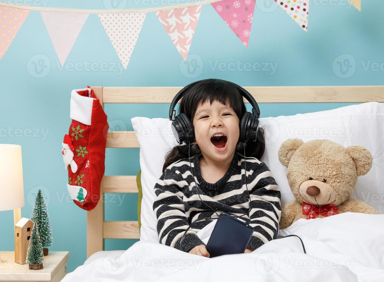 söt liten flicka sång med smartphone med teddy Björn i henne sovrum, Lycklig asiatisk barn liten flicka lyssnande de musik med hörlurar med teddy Björn på de säng, teknologi begrepp foto