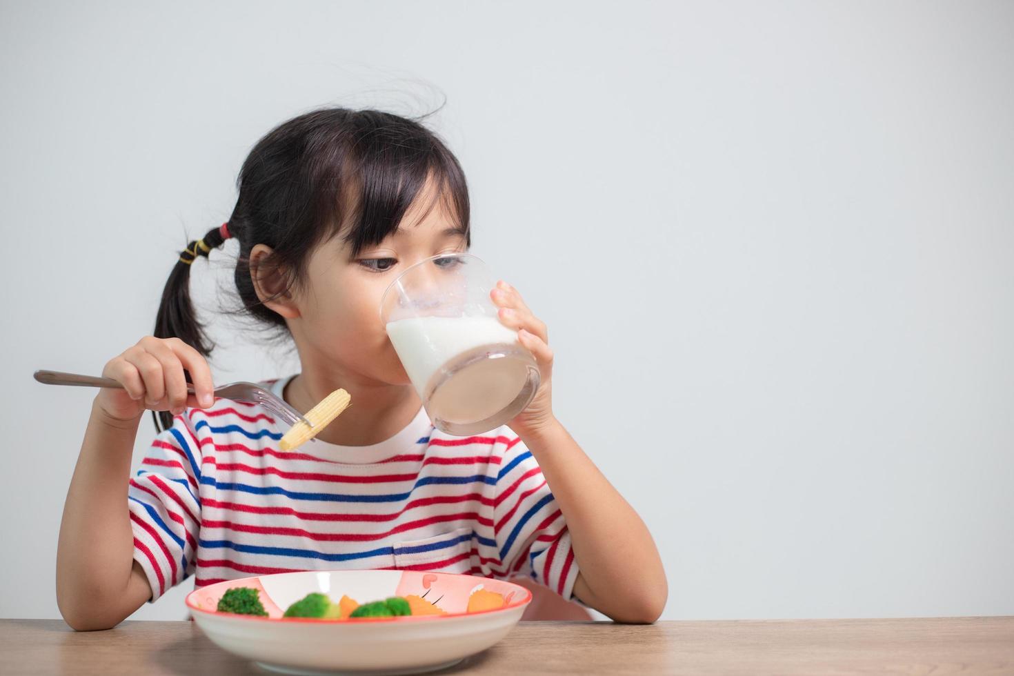 söt asiatisk barn flicka äter friska grönsaker och mjölk för henne måltid foto