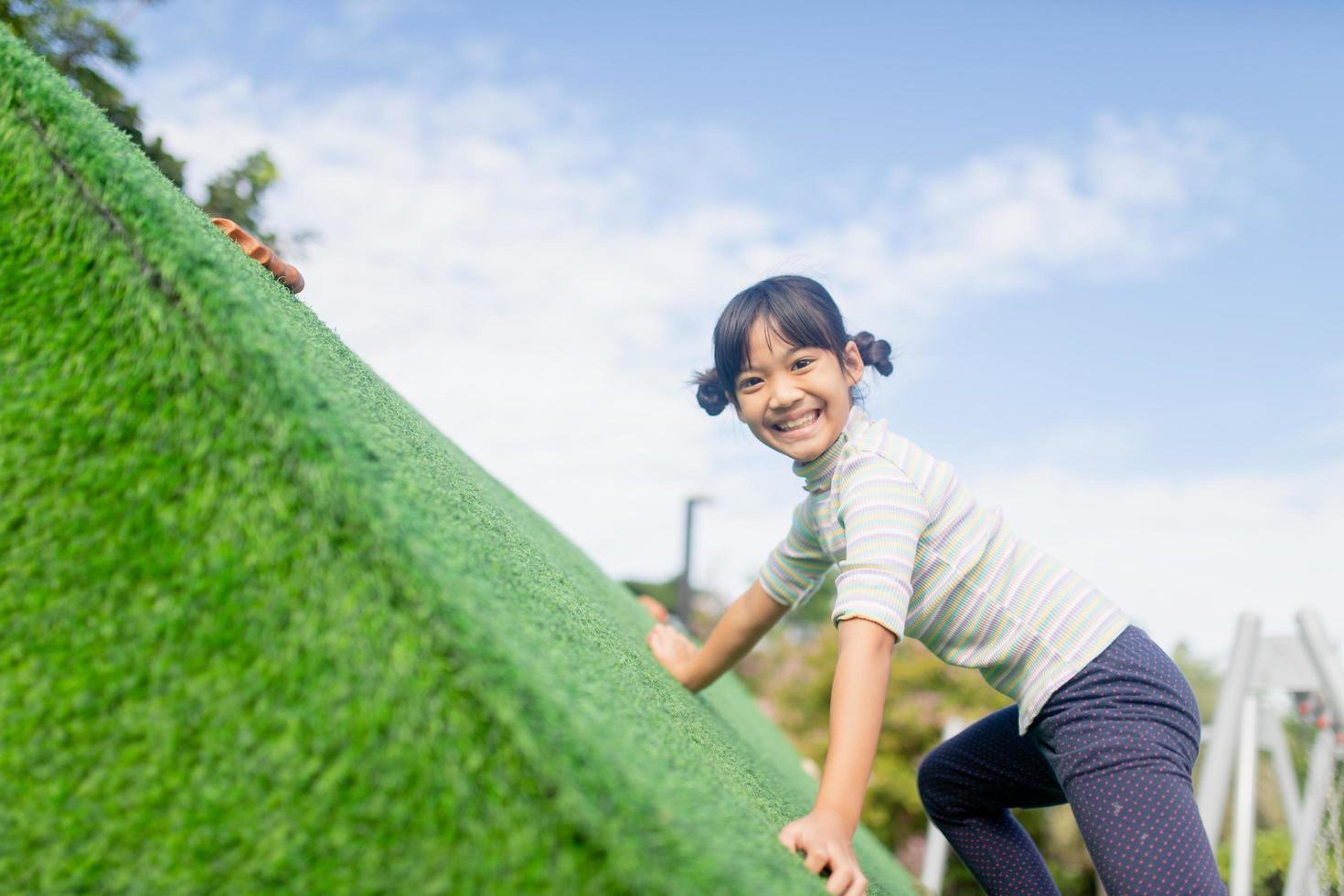 söt asiatisk tjej som har roligt försöker klättra på konstgjorda stenblock på skolgårdens lekplats, liten flicka som klättrar uppför bergväggen, hand-öga-koordination, kompetensutveckling foto