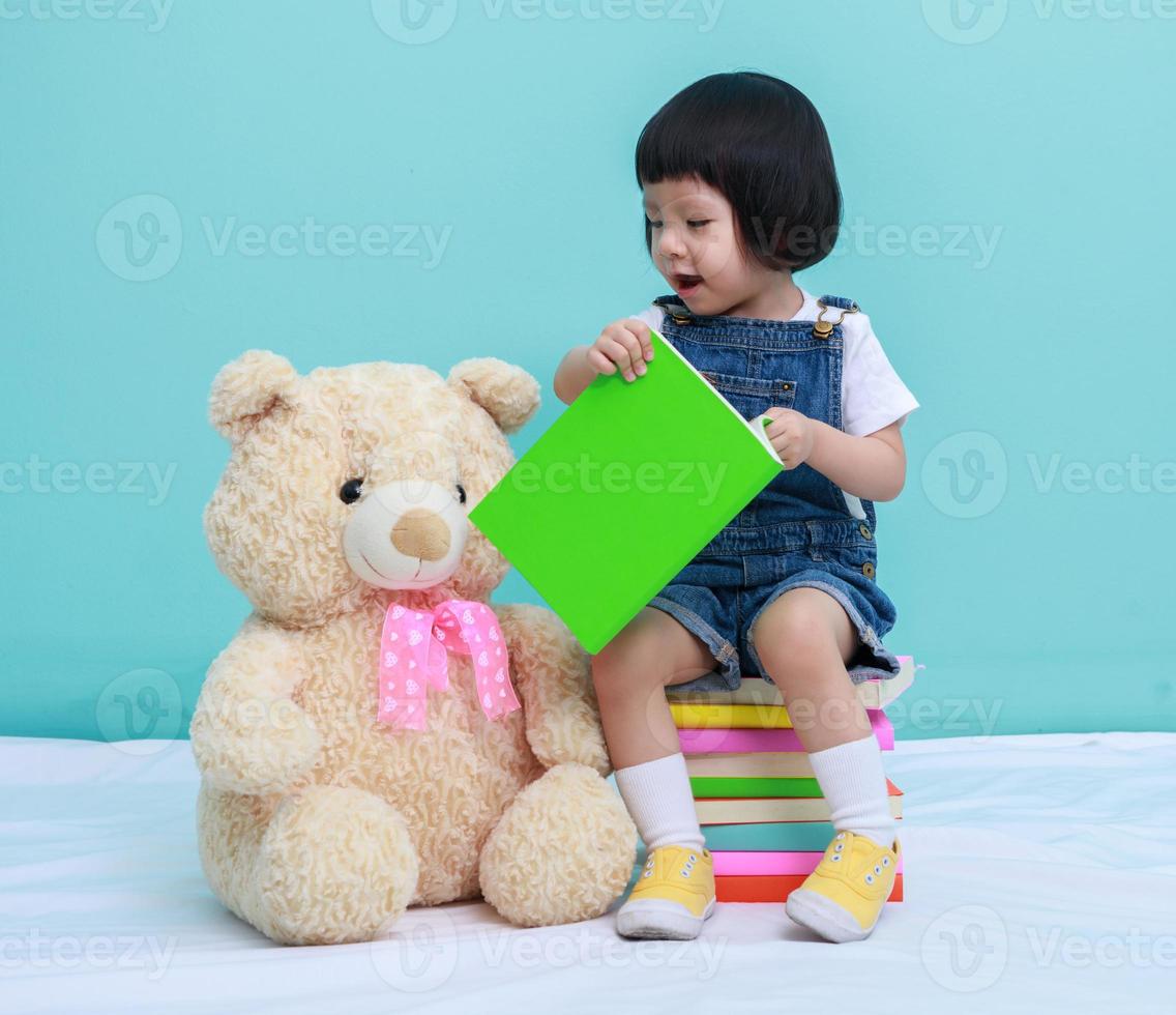 barn liten asiatisk flicka eller en liten söt asiatisk flicka läsning en bok i de grön bakgrund och Sammanträde på de böcker med en leksak teddy Björn foto