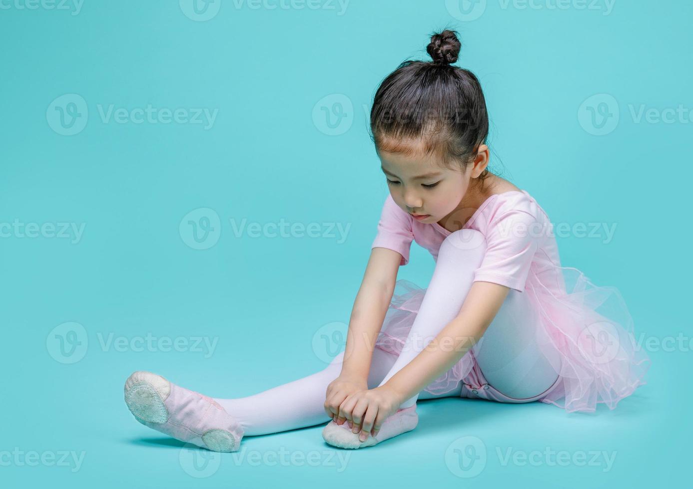 skön leende asiatisk liten flicka i en rosa kostym är dans en balett på skola, tömma Plats i studio skott isolerat på färgrik blå bakgrund foto