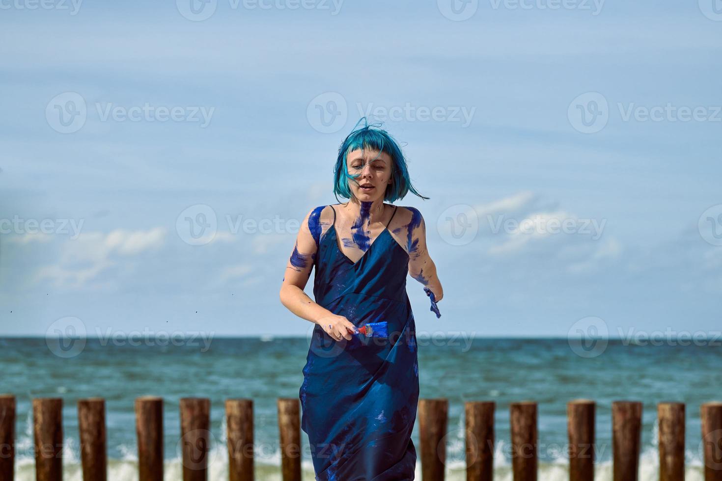 konstnärlig blåhårig kvinna performance artist insmord med blå gouache färger dans på stranden foto